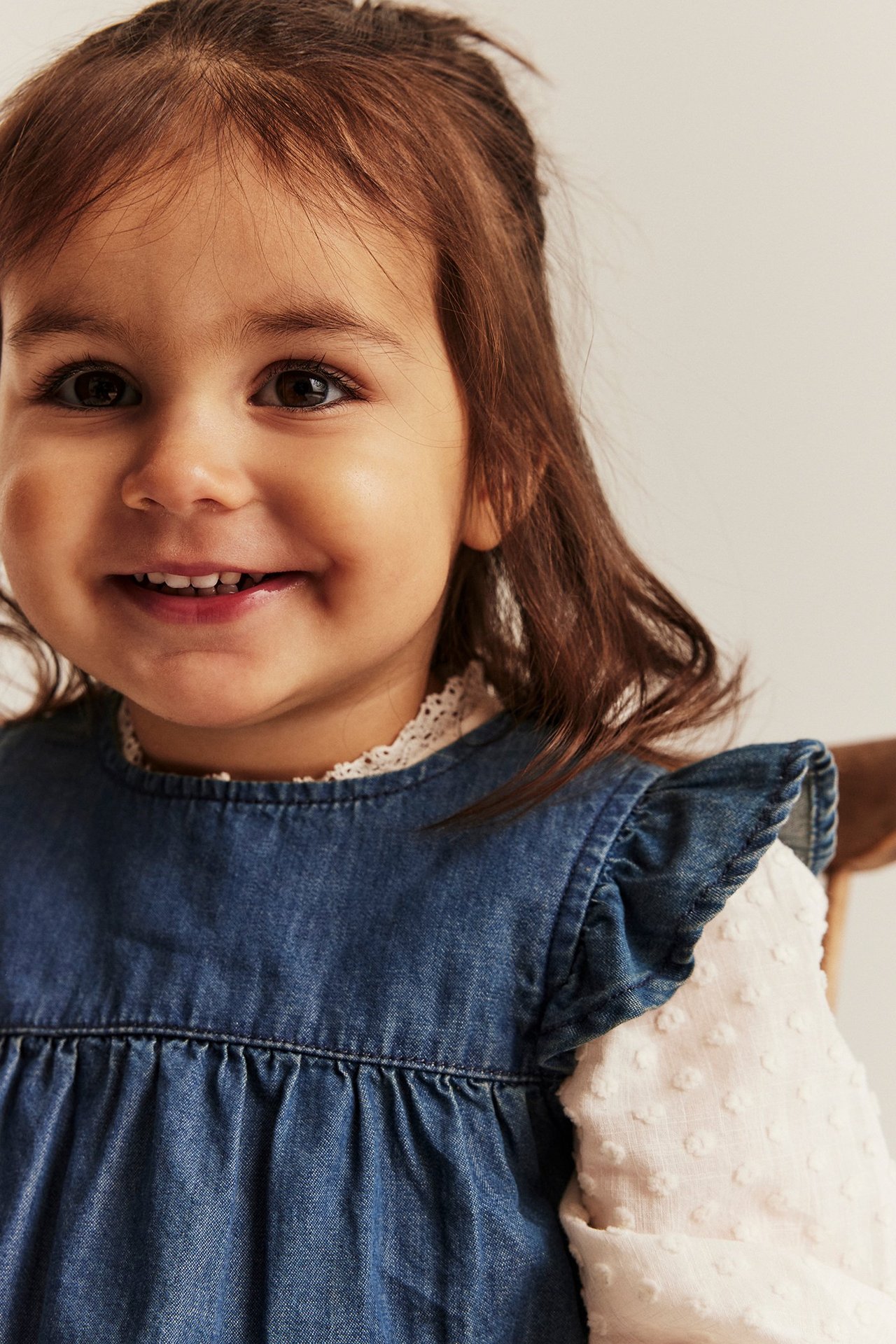 Close-up of a smiling baby girl wearing a blue denim ruffled dress over a white textured long-sleeved top.