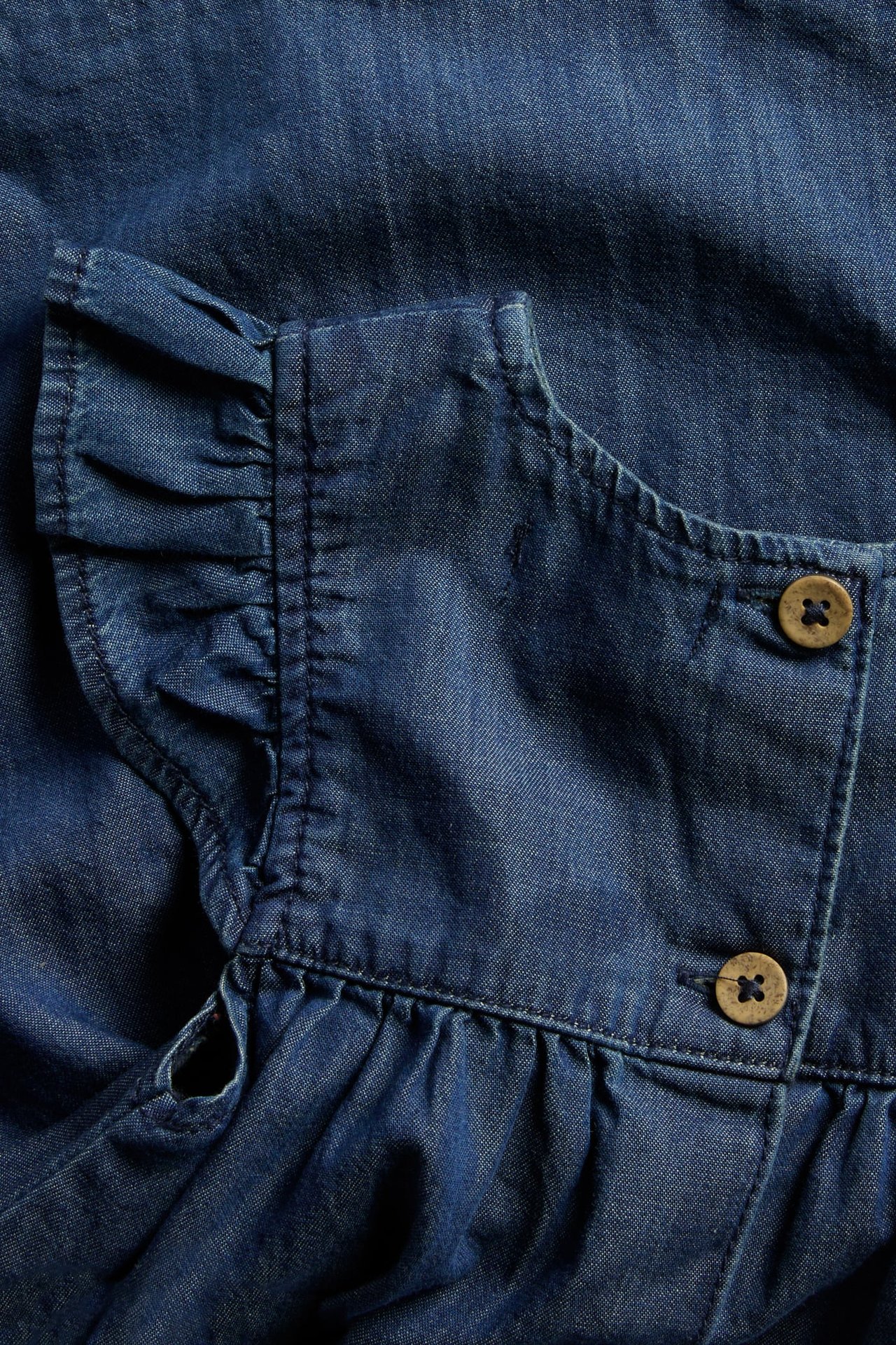 Close-up of a baby's denim dress with ruffled shoulders and back fastening with two brown buttons.