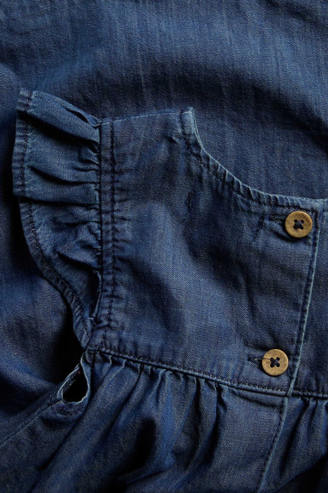 Nahaufnahme eines Baby-Jeanskleides mit Rüschen an den Schultern und zwei braunen Knöpfen am Rücken.