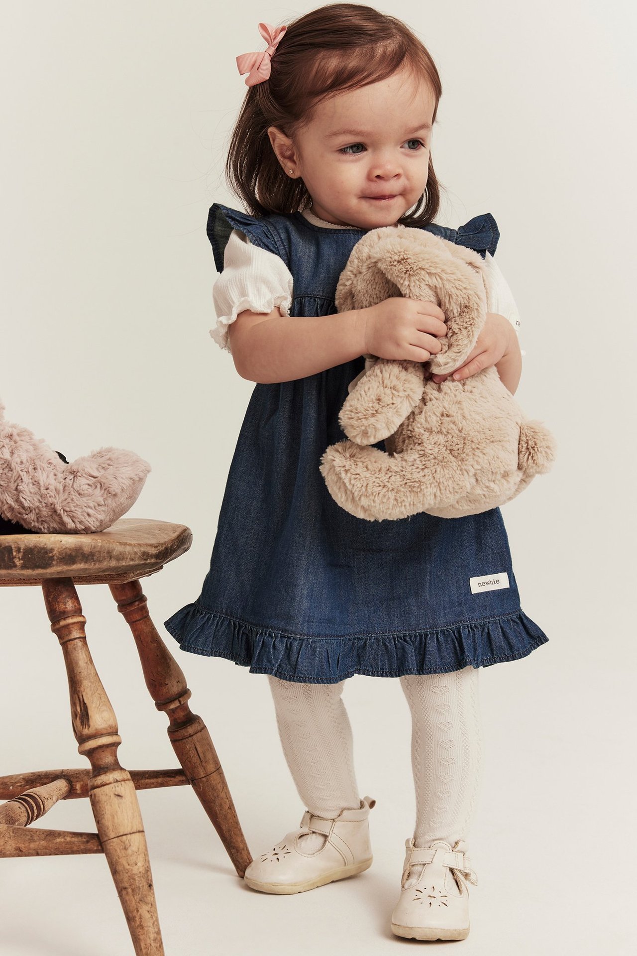 Baby trägt blaues Jeans-Rüschenkleid über weißem Oberteil, gemusterter Strumpfhose und beigen Schuhen.