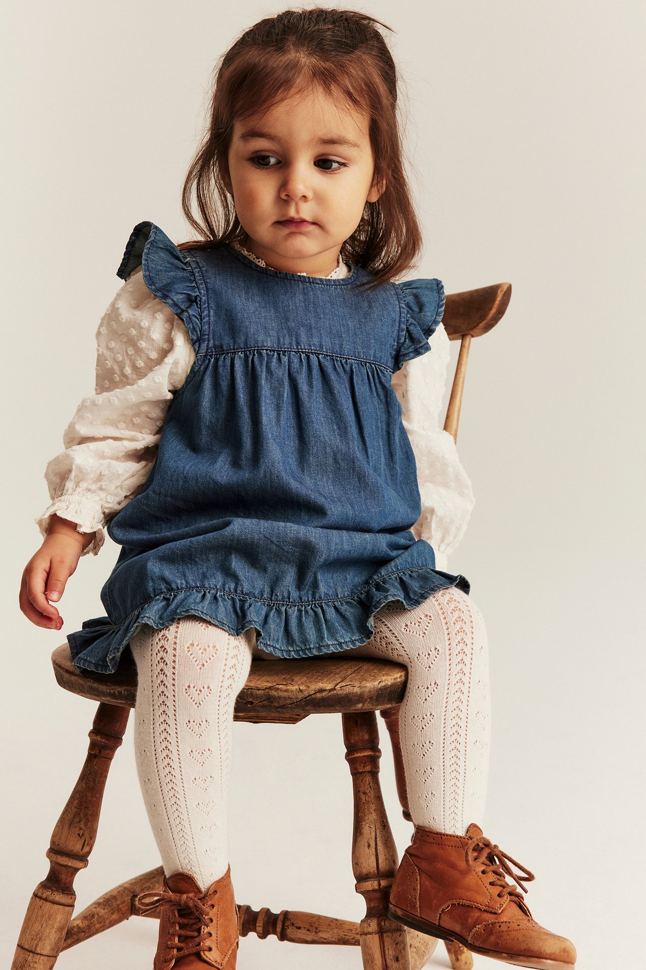 Front view of a baby wearing a denim ruffle dress, white textured shirt, white heart tights, and brown boots.