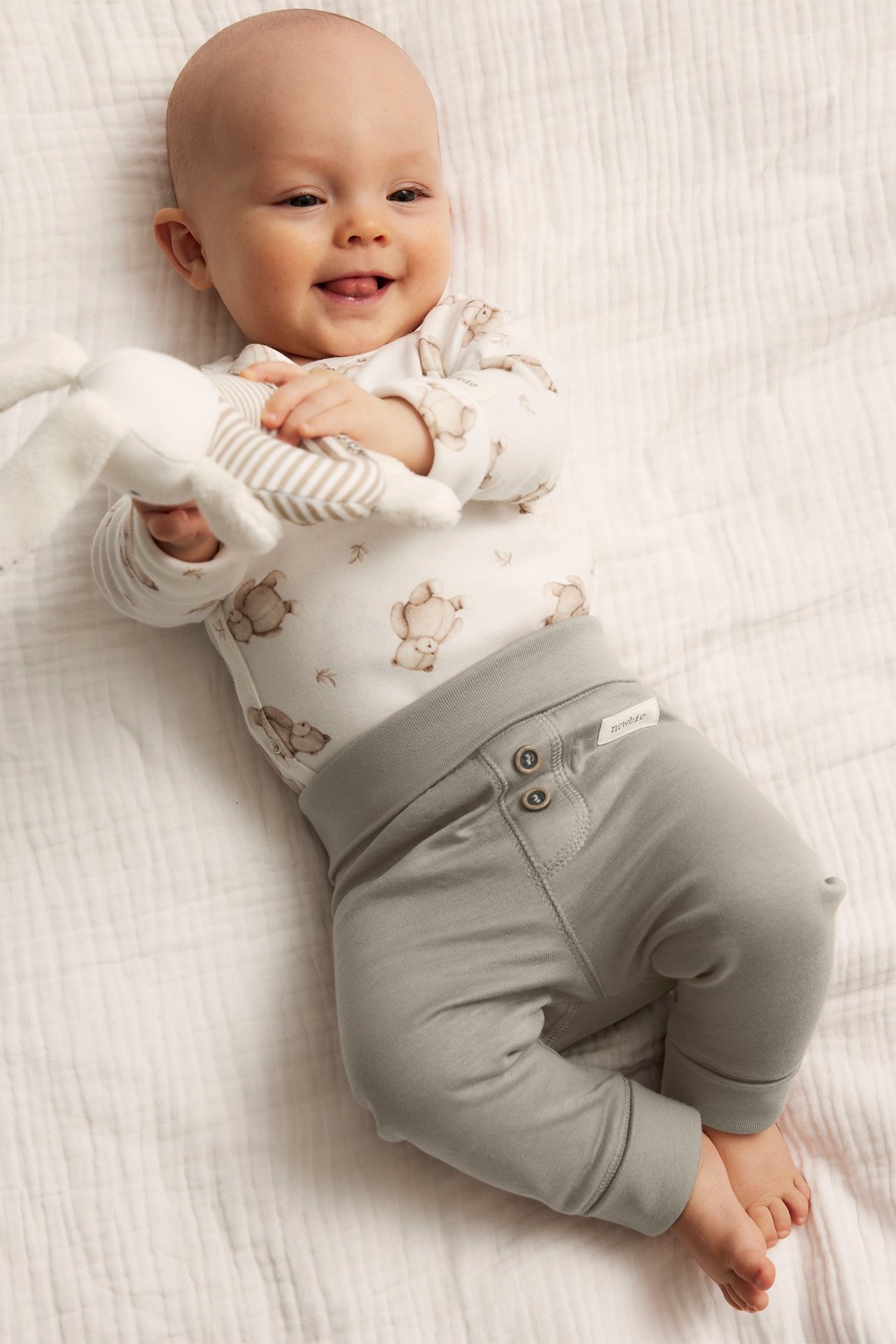 Top-down view: Baby in light grey cotton leggings with buttons and a white teddy bear print bodysuit, holding a toy.
