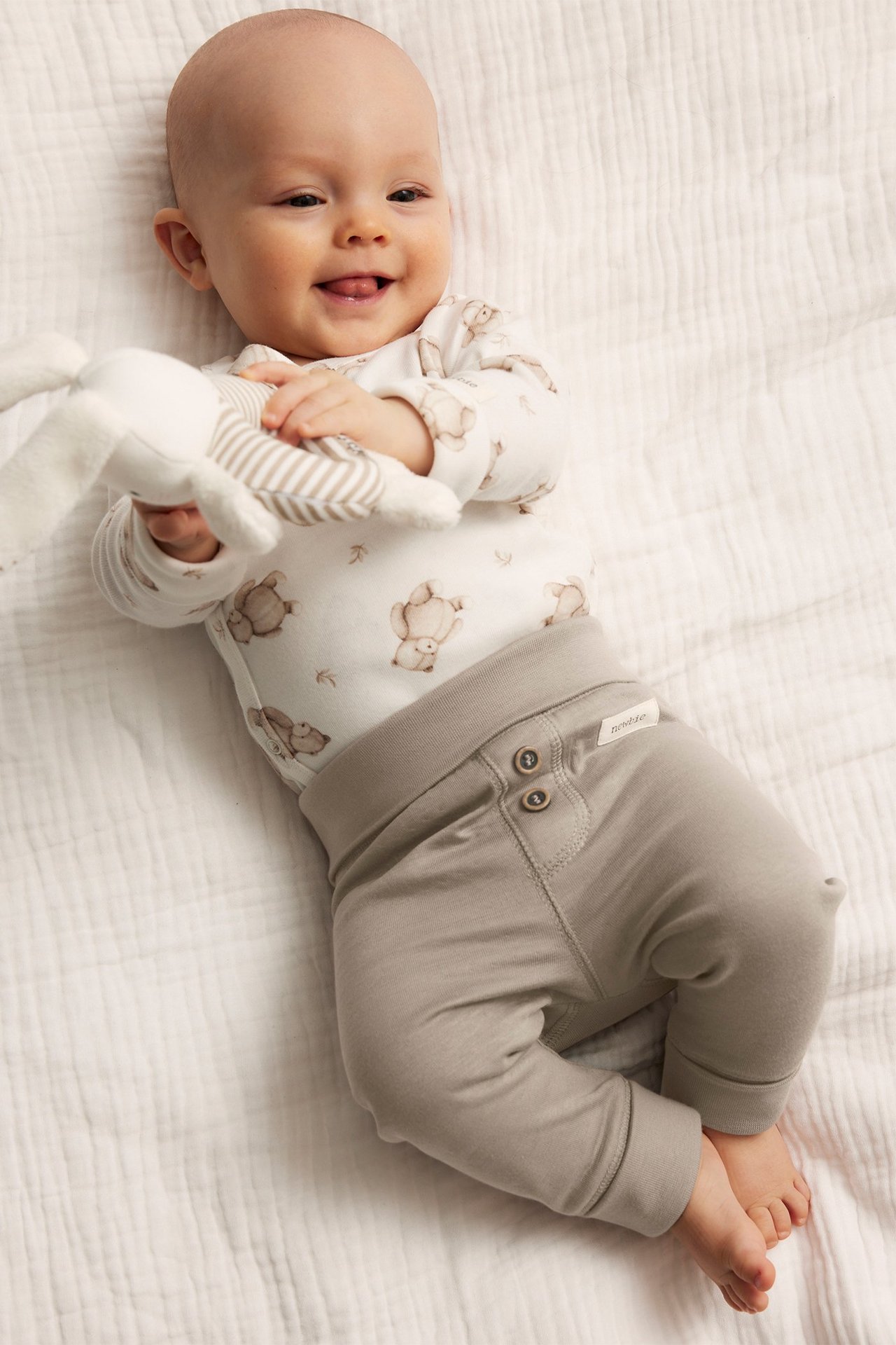 Smiling baby wears beige cotton leggings with buttons and a white teddy print bodysuit, holding a plush toy.