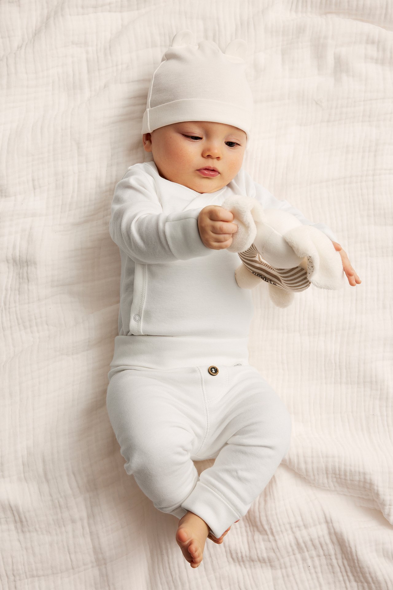 Top view: Baby in off-white cotton leggings with buttons, matching wrap top, and hat, holding a plush toy.