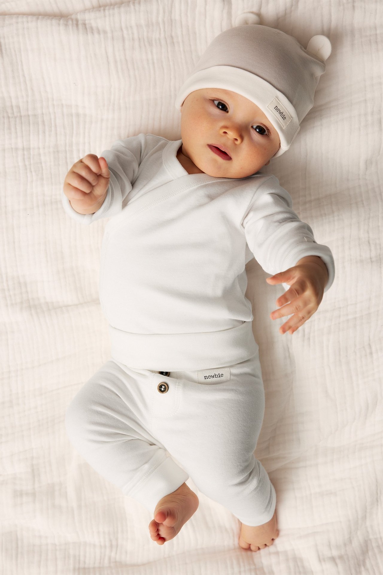 Vorderansicht eines Babys in cremeweißen Baumwoll-Leggings mit Knöpfen, weißem Wickeloberteil und grauer Ohrenmütze.