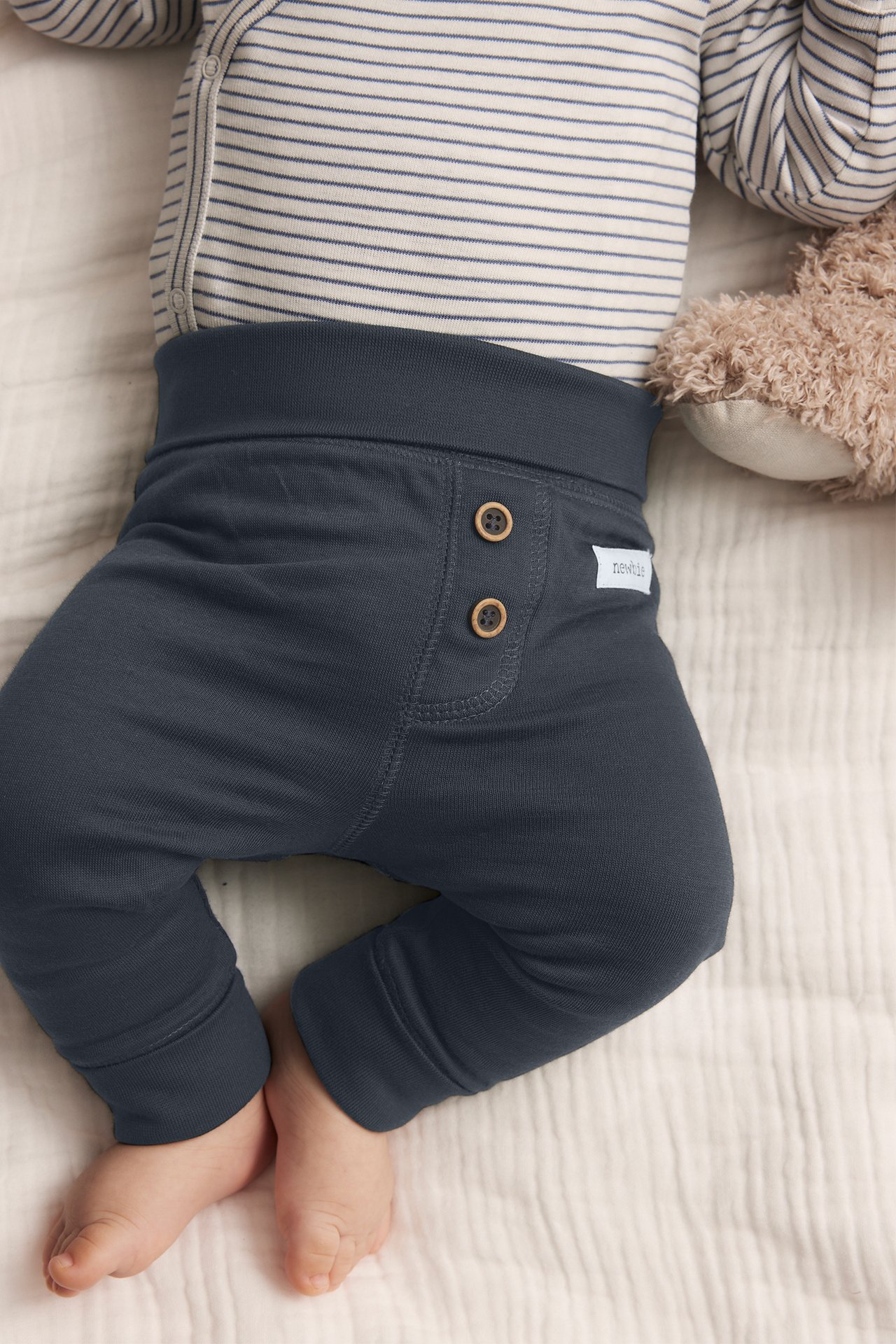 Draufsicht auf ein Baby in dunkelblauen Baumwoll-Leggings mit Holzknöpfen und einem gestreiften Body.
