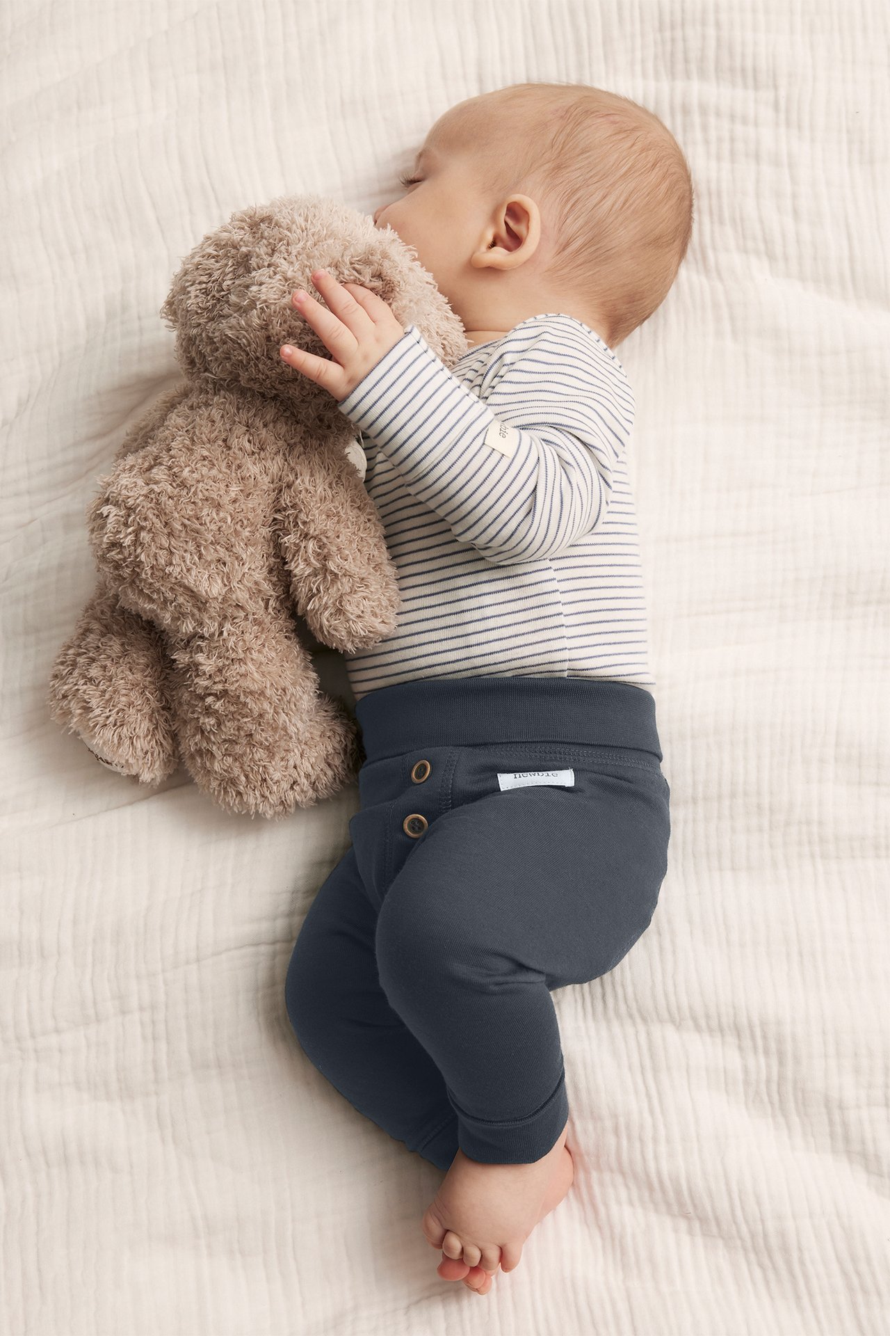 Draufsicht: Baby in dunkelblauen Baumwoll-Leggings mit Knöpfen, gestreiftem Body, kuschelt Teddybär.