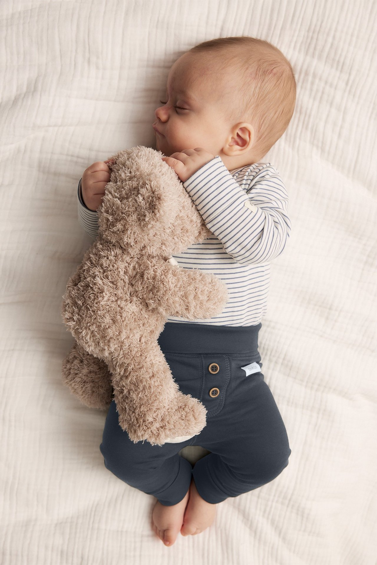 Overhead view of a sleeping baby in dark blue buttoned leggings, a striped bodysuit, holding a brown teddy bear.