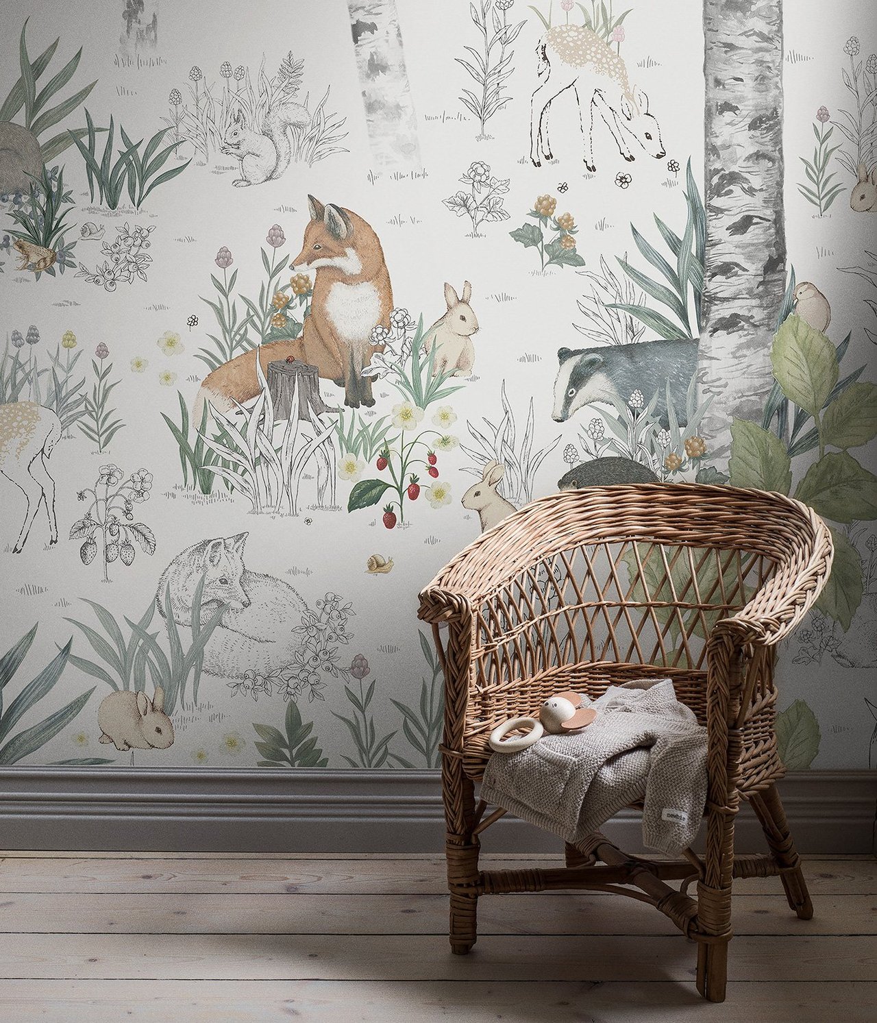 Vorderansicht: Helle Tapete mit Waldtieren und Birken. Korbstuhl hält eine graue Decke und Holzrassel.