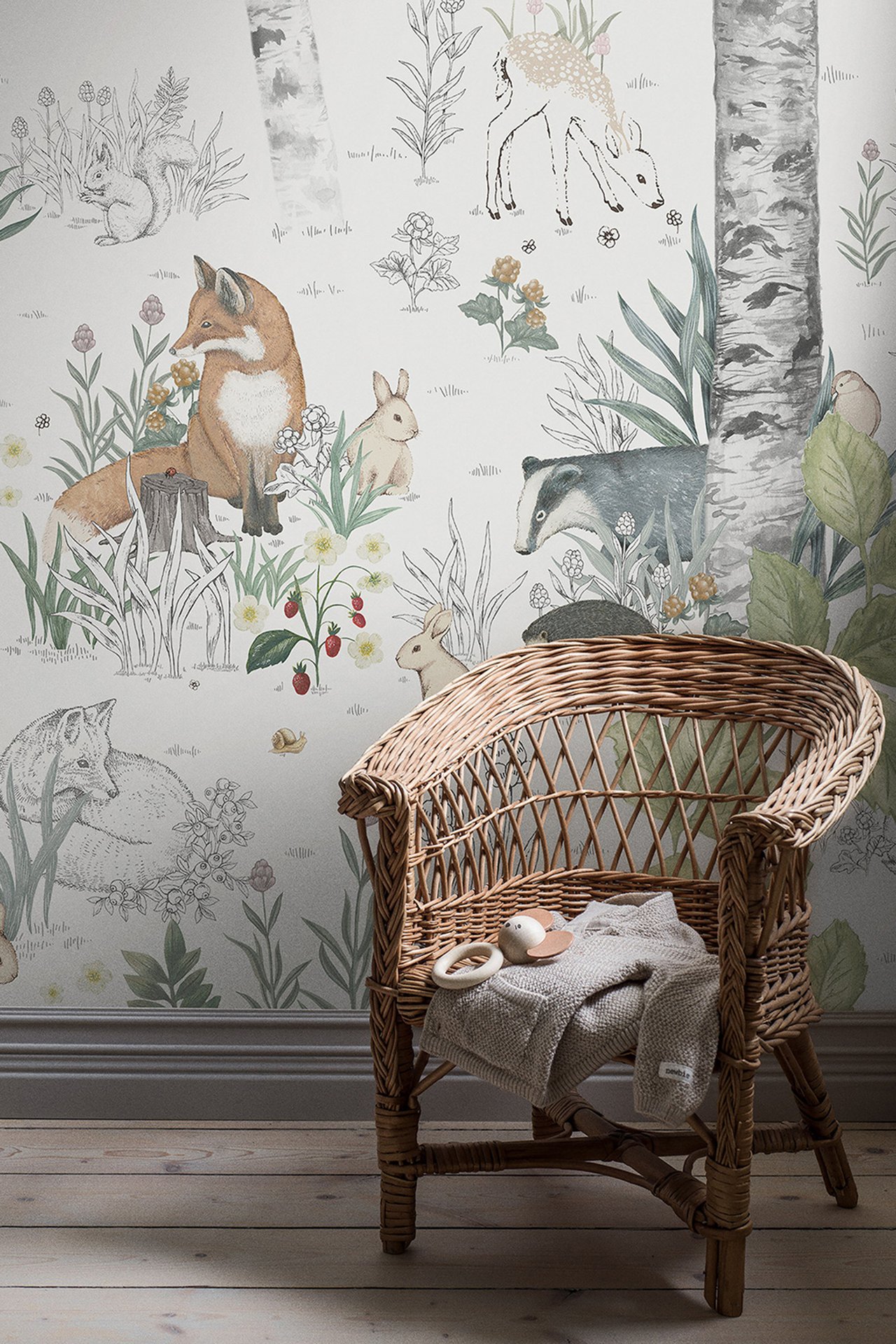Front view: Light wallpaper with forest animals and plants. A wicker chair holds a beige knit blanket and toy.