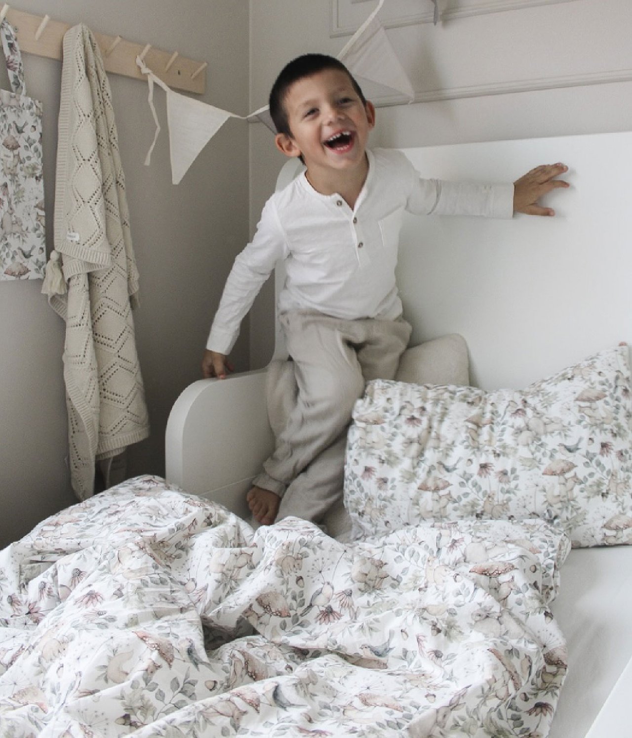 Front view: Light beige bed set with forest print. A kid laughs on bed. Blanket and bag hang.