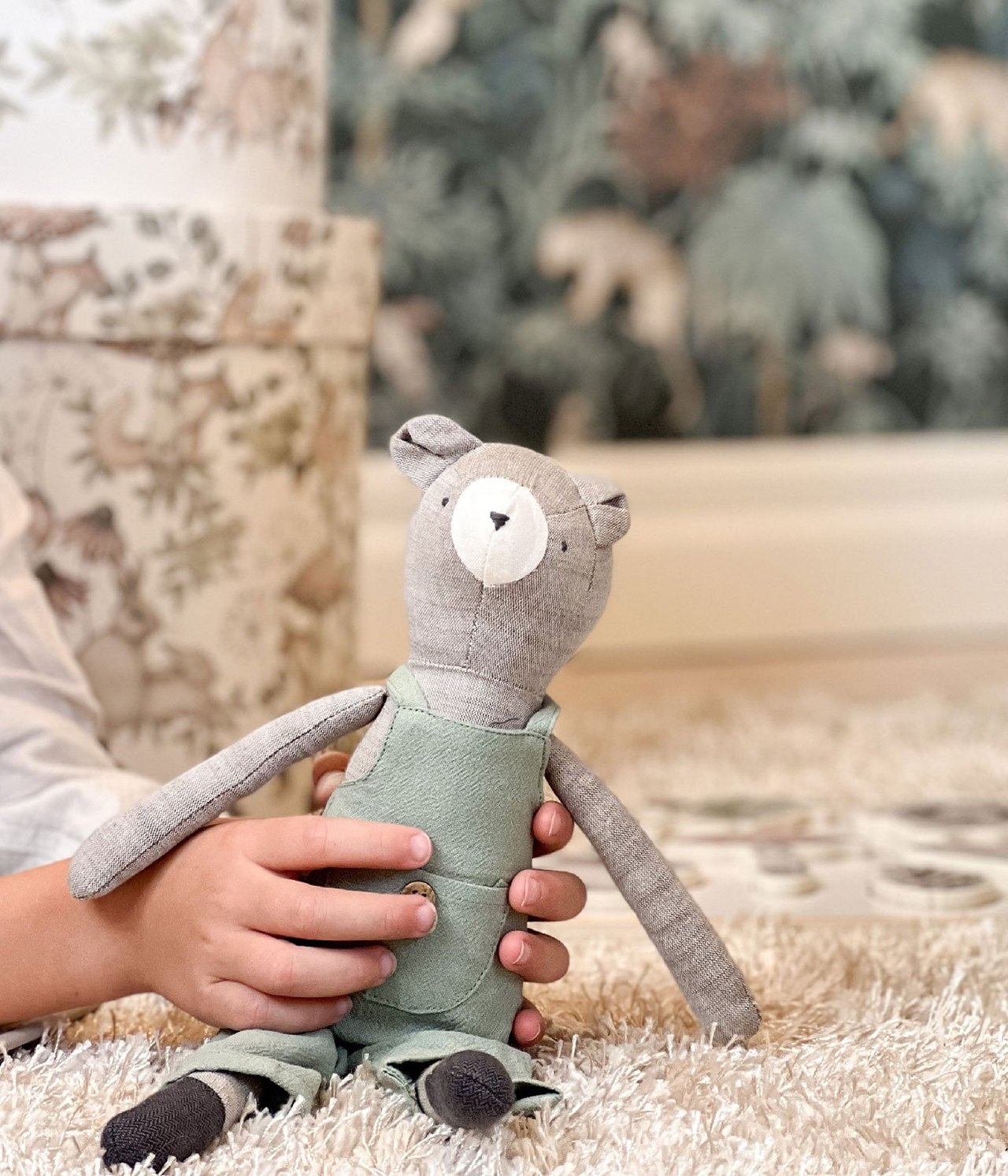 Nahaufnahme von Kinderhänden, die einen grauen Stoffbären mit grüner Latzhose halten.