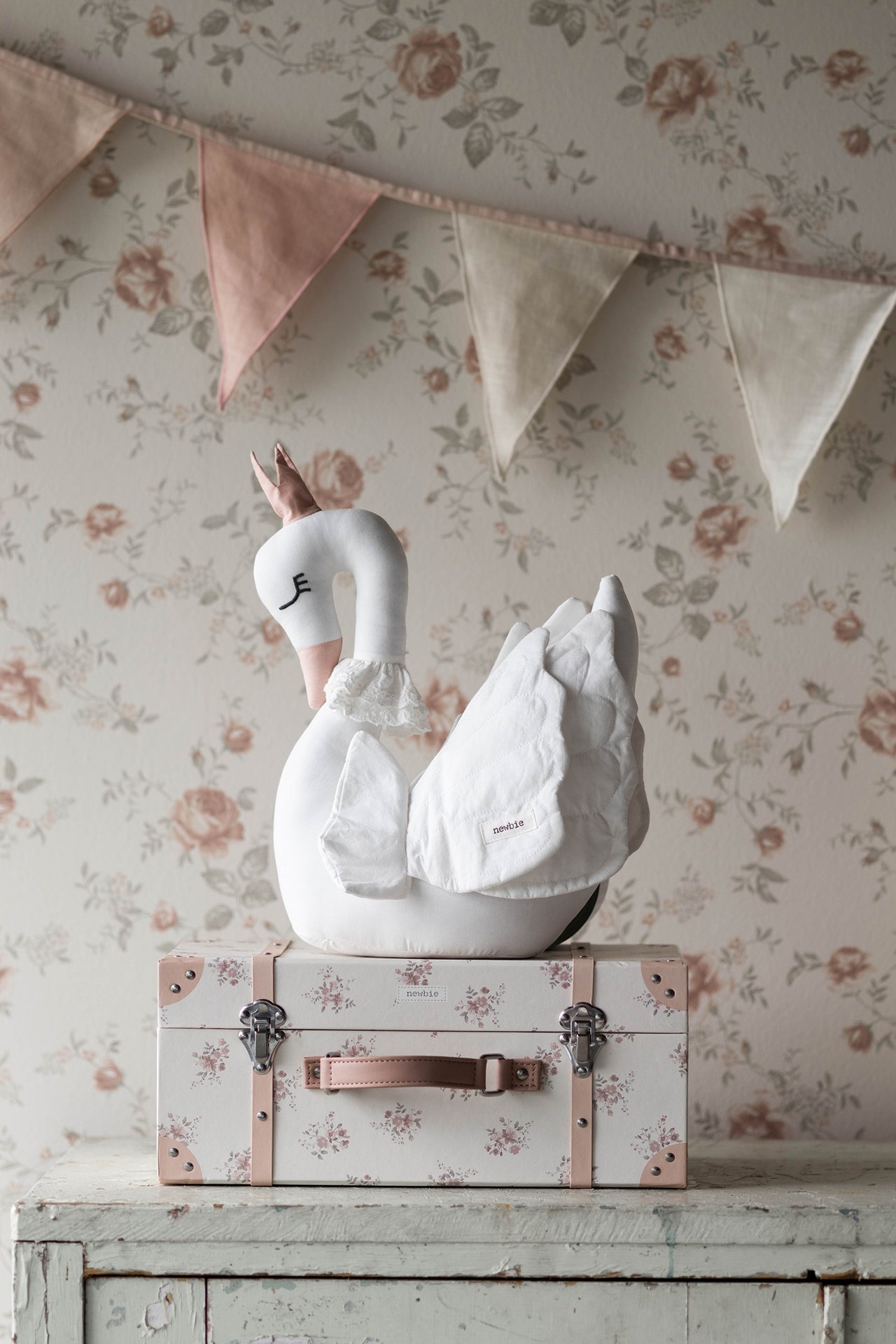 Front view: Off-white cotton swan soft toy with pink crown, ruffled collar, on a floral trunk.