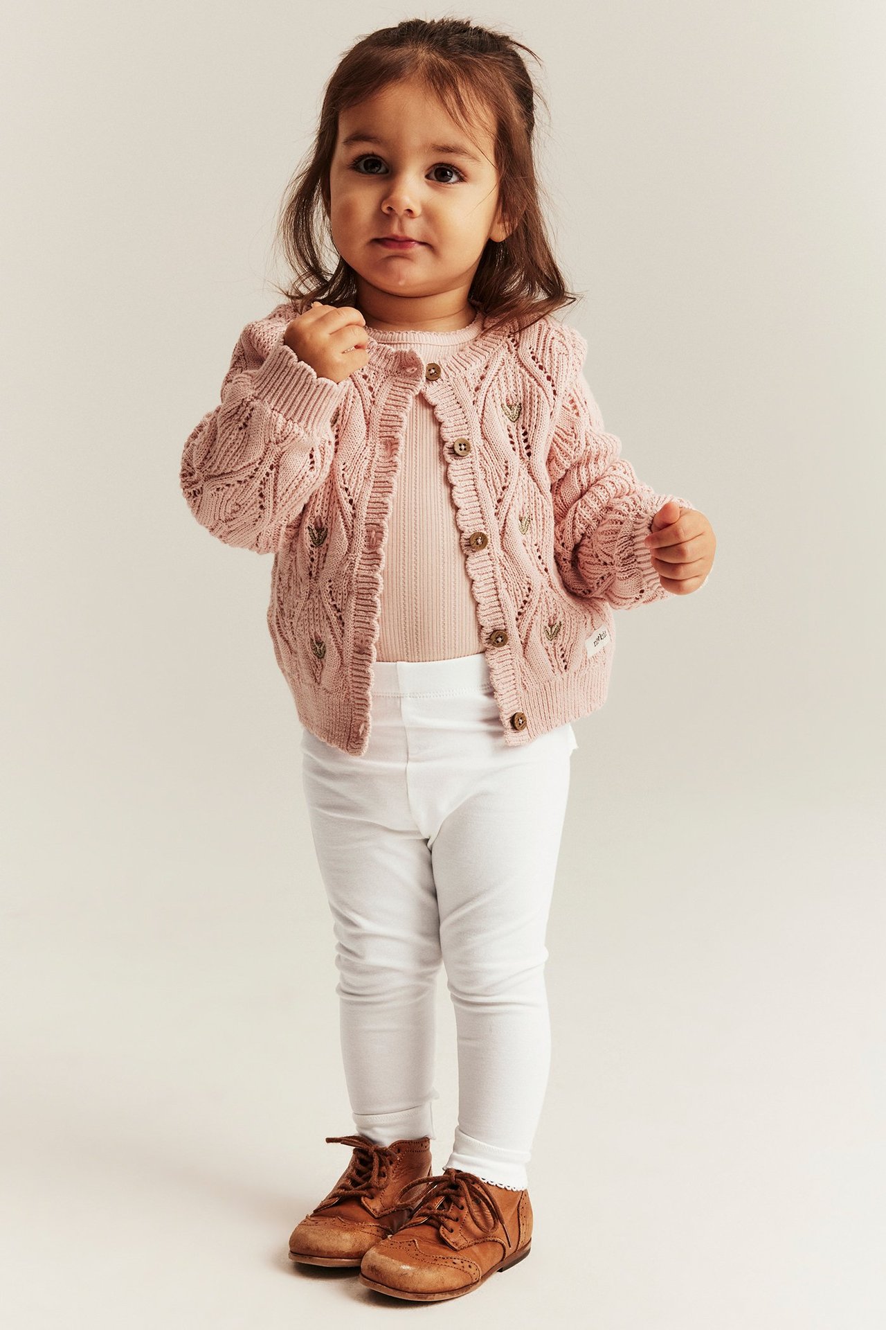 Front view of a baby in offwhite leggings, a pink knitted cardigan, pink top, and brown shoes.