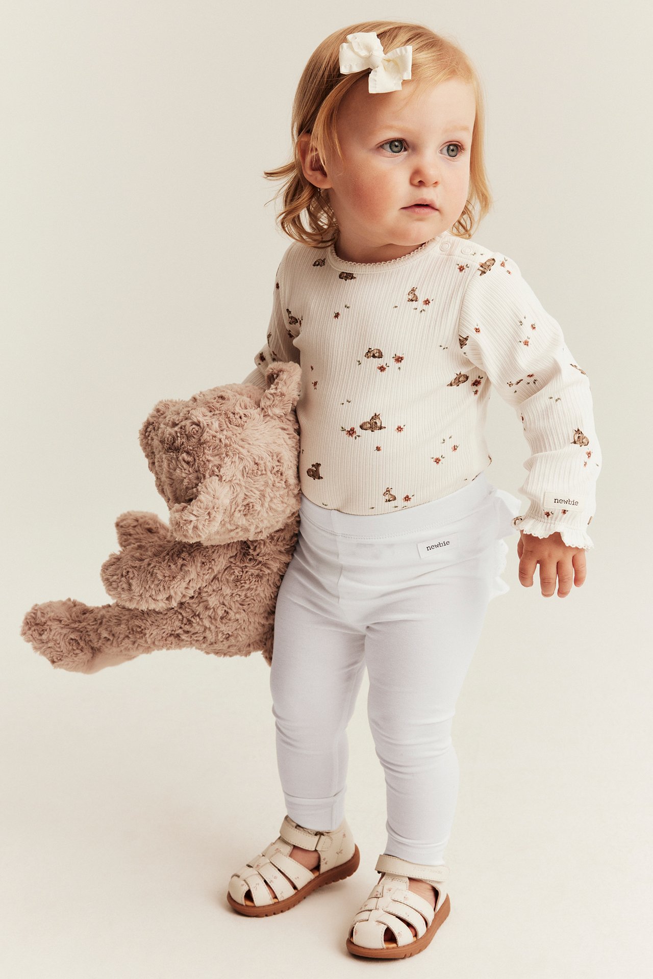 Baby trägt weißen Body mit Rehmuster, weiße Leggings und Sandalen, hält einen Teddybär.