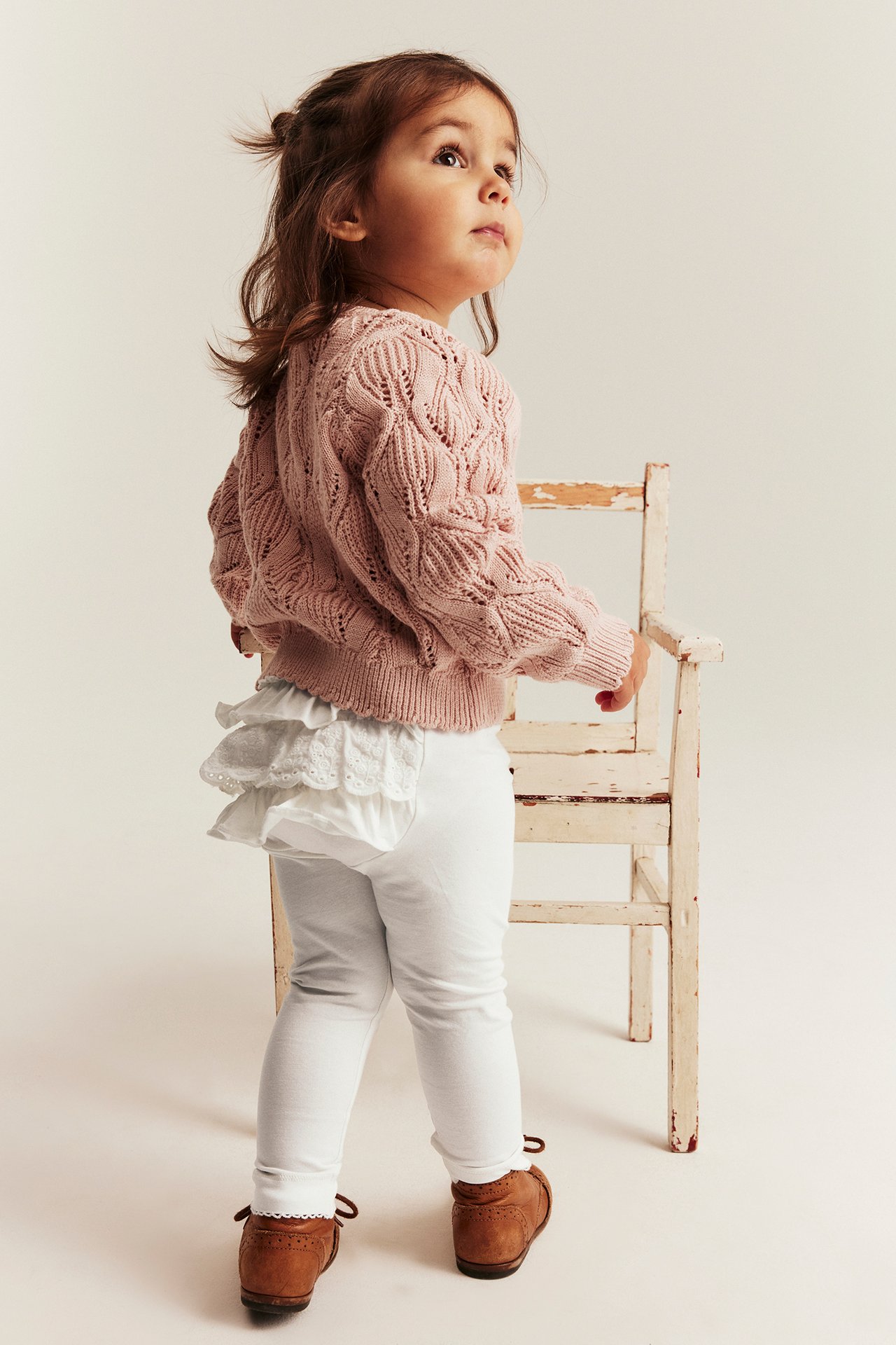 Baby von hinten in cremeweißen Rüschen-Leggings, rosa Strickpullover und braunen Schuhen.