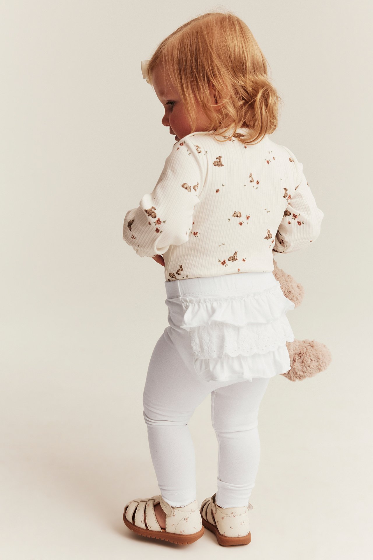 Rückenansicht eines Babys in weißem gerippten Hasen-Langarmtop, weißen Rüschenleggings und beigen Sandalen.