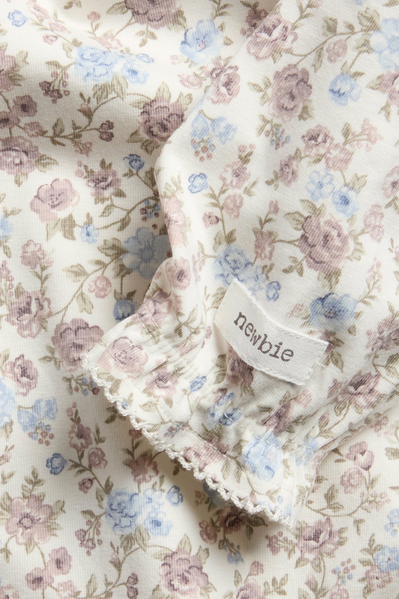 Close-up of an off-white cotton blend baby bodysuit with a floral pattern, ruffle, and picot trim.