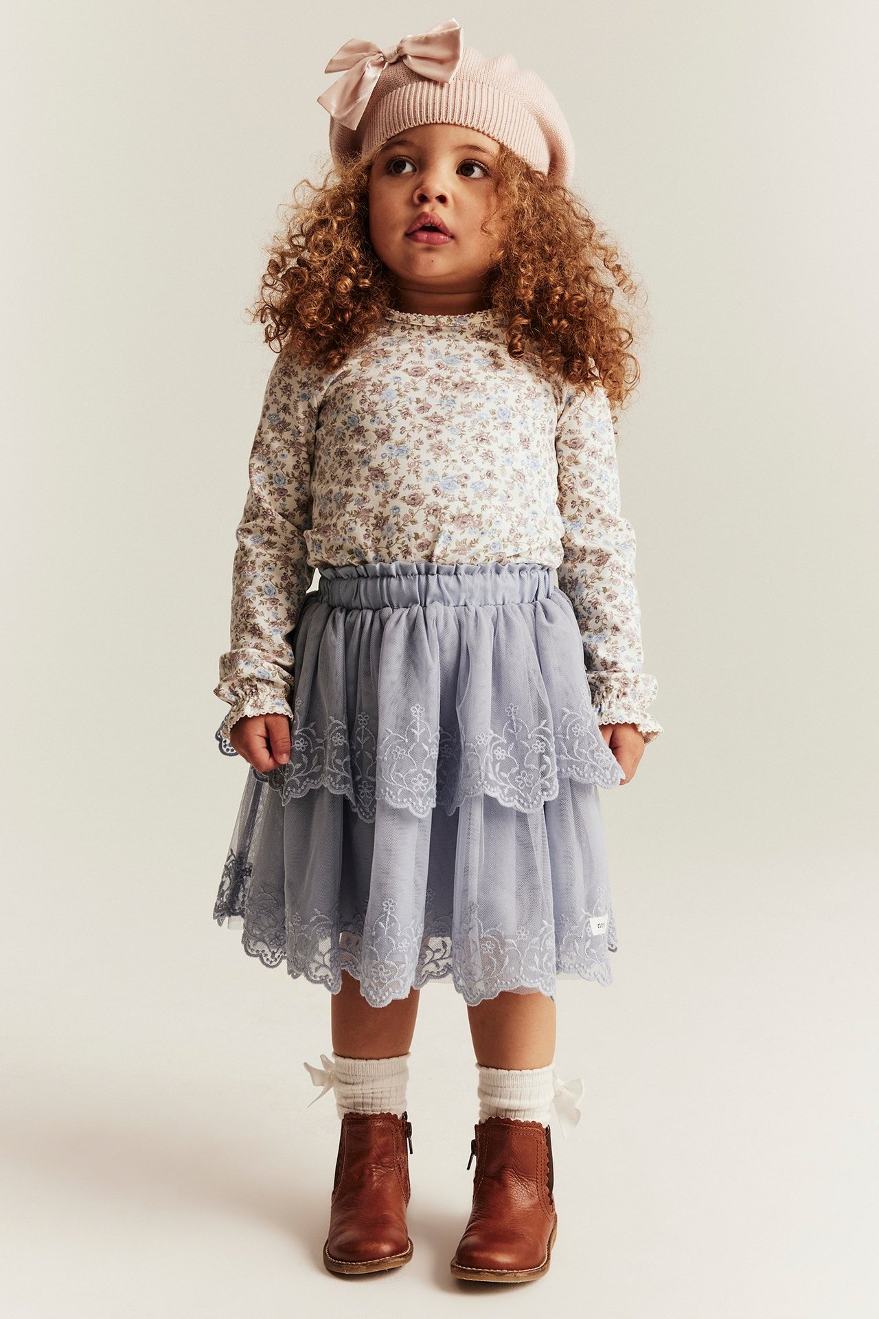 Front view: kid in off-white floral long-sleeve top, blue tiered skirt, pink beret, white socks, brown boots.