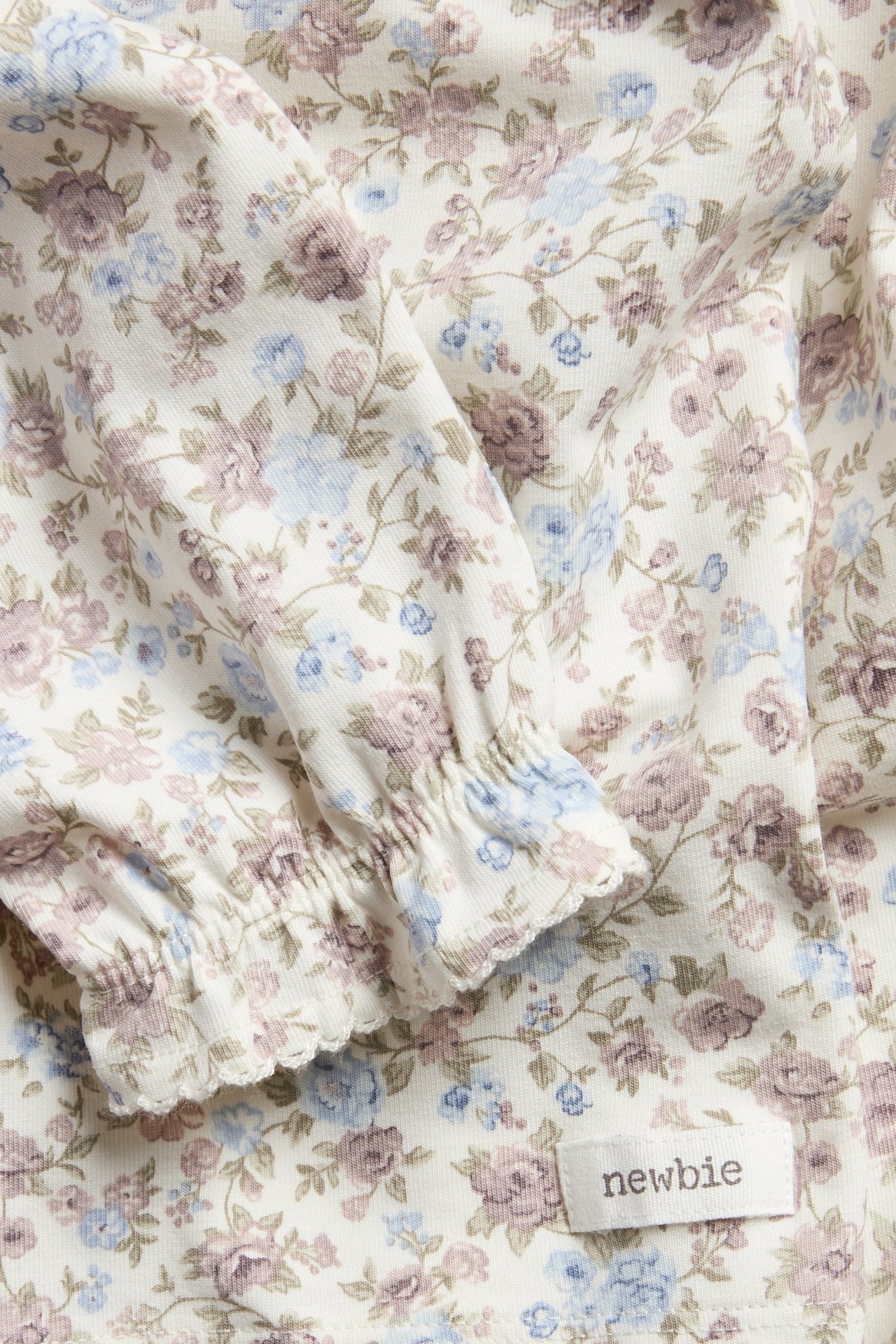 Close-up: off-white floral long-sleeved top with light blue and purple flowers, ruffled cuffs, and a brand label.