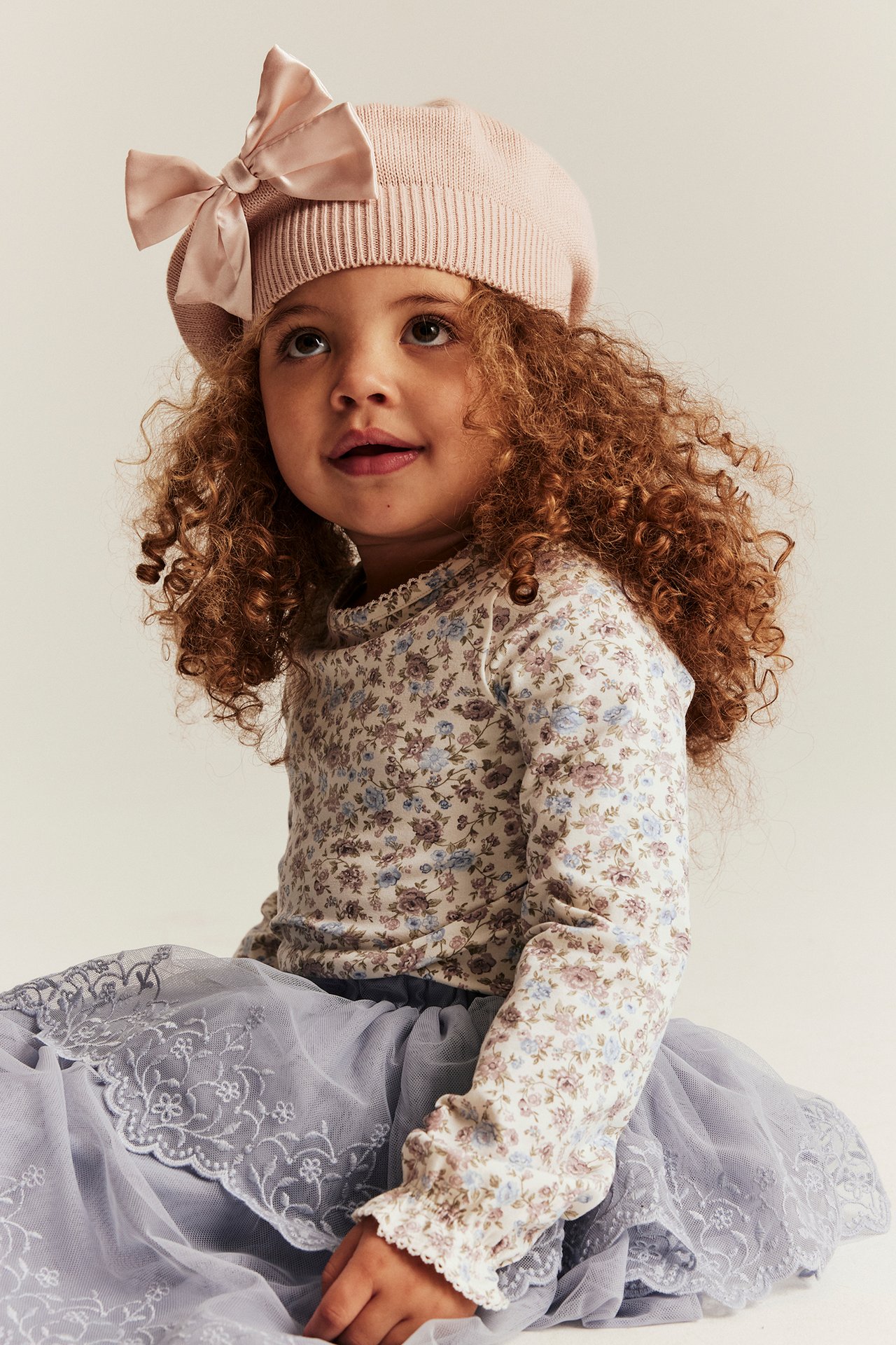 Kid in off-white floral long-sleeved top with picot trim, pink bow beret, and grey tulle skirt.