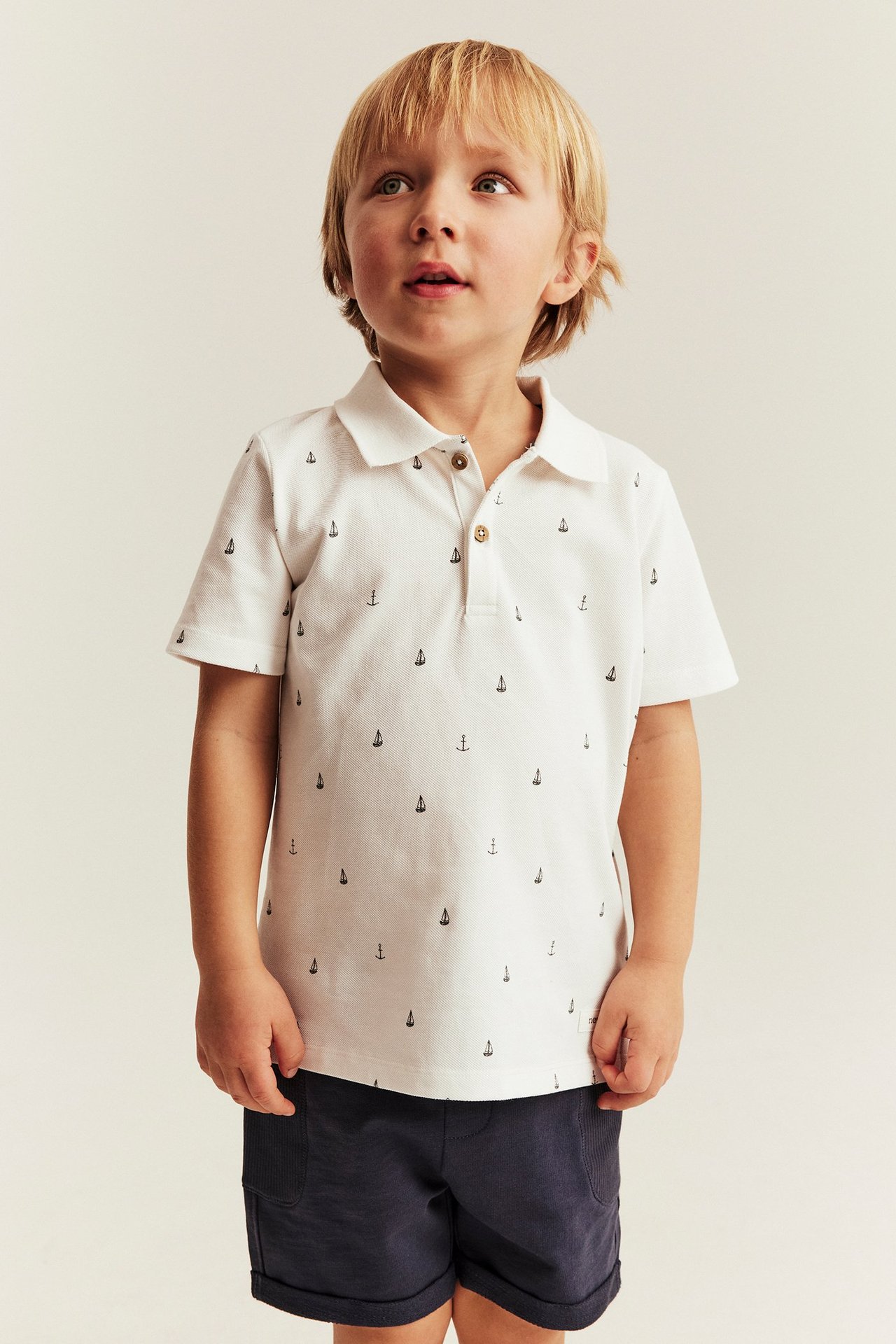 Front view of a boy in an off-white cotton polo shirt with marine pattern and dark blue shorts.