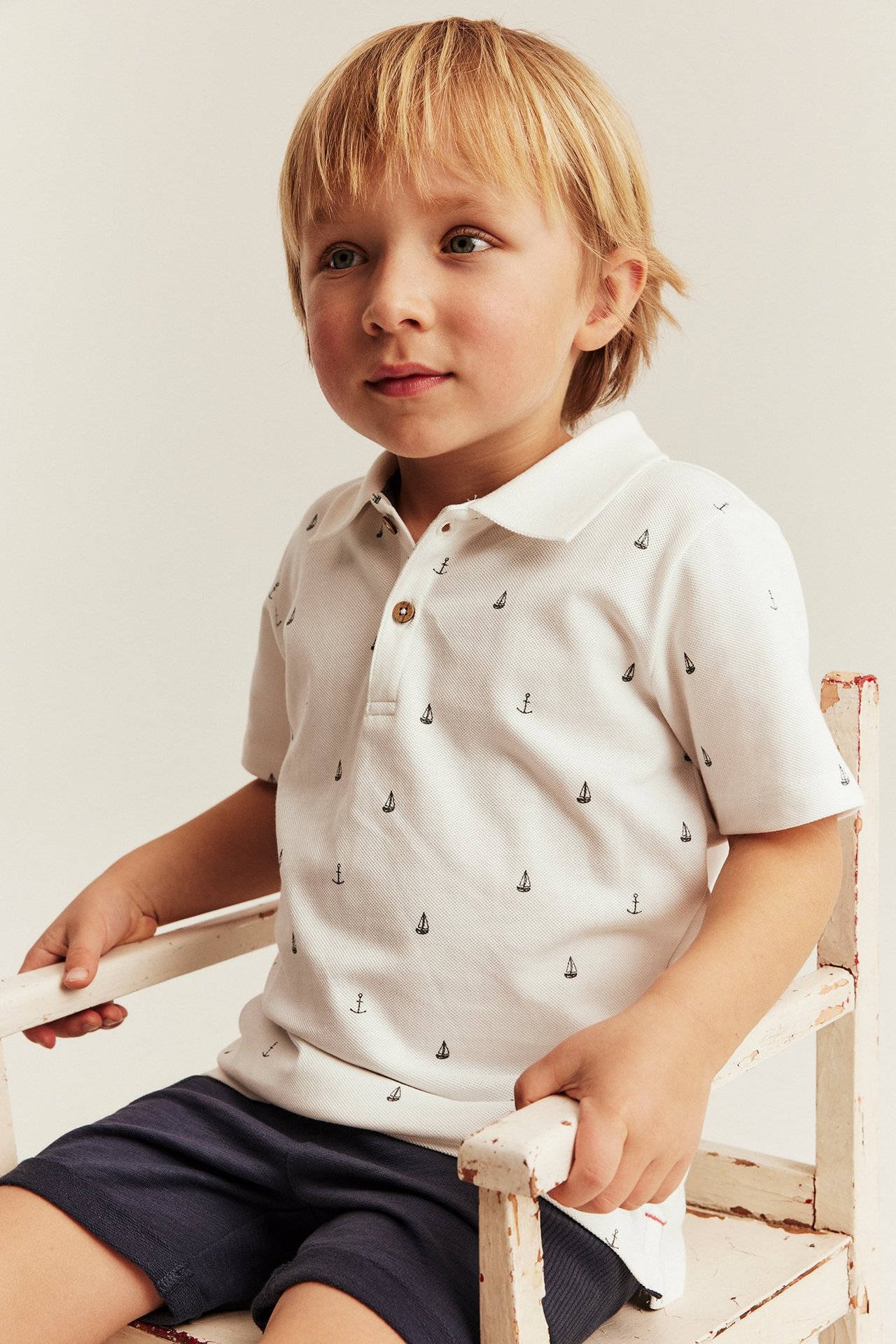 Front view of a kid wearing an off-white cotton polo shirt with a marine print and dark blue shorts.