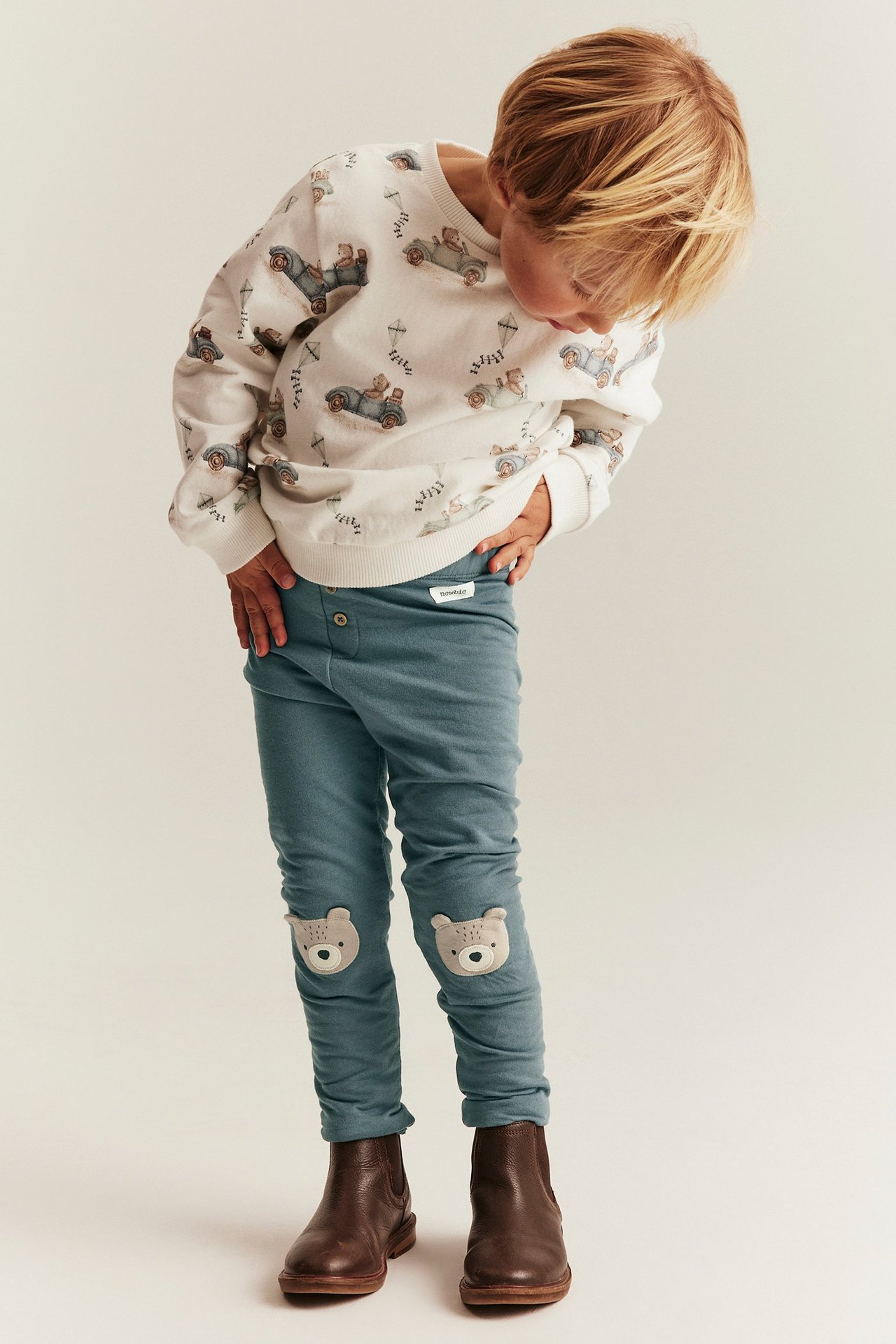 Frontansicht: Kind in blauen Leggings mit Bären-Knieapplikationen, weiß gemustertem Sweatshirt und braunen Stiefeln.