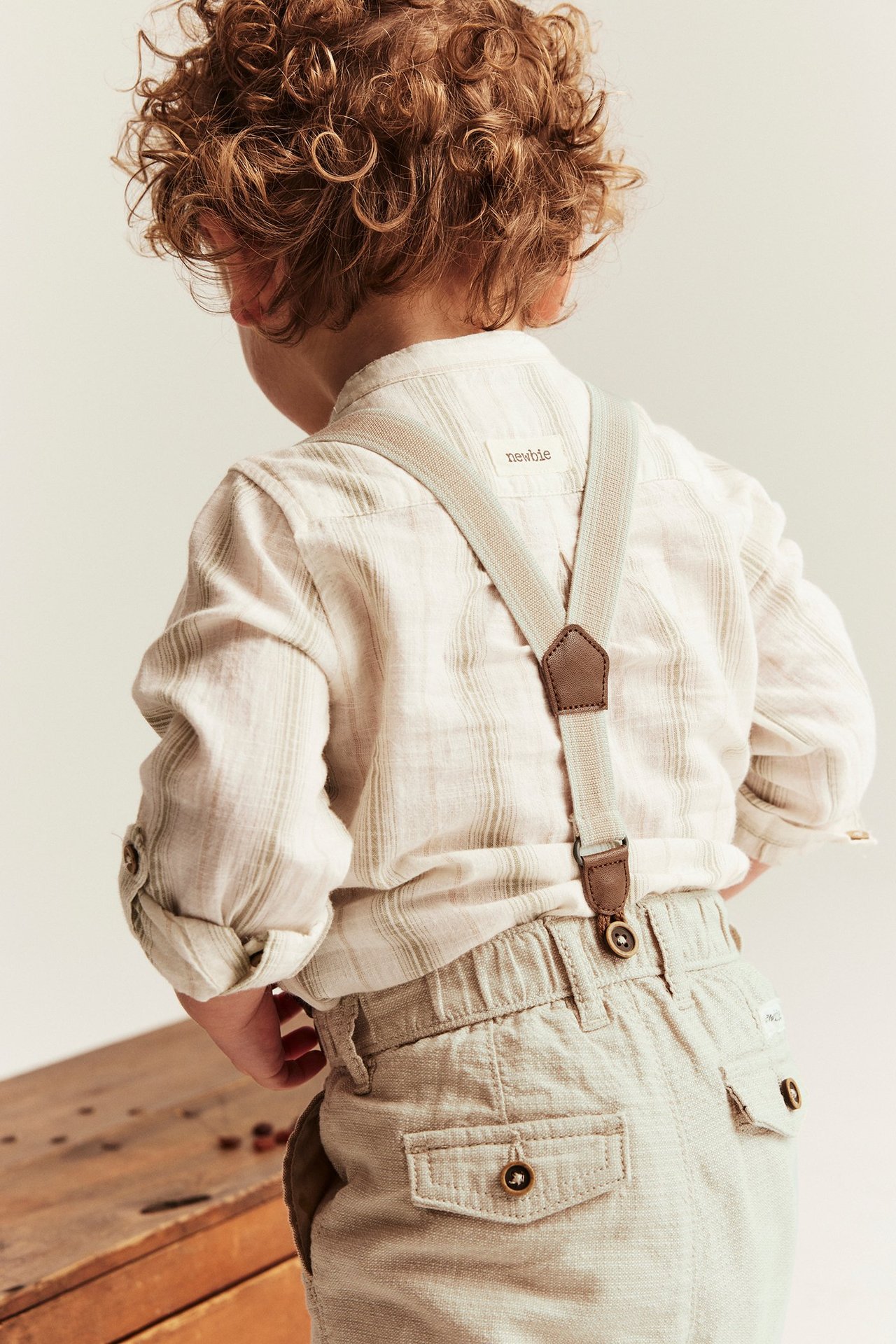 Kid's back view: beige woven suspender pants with faux leather details worn over a striped shirt.