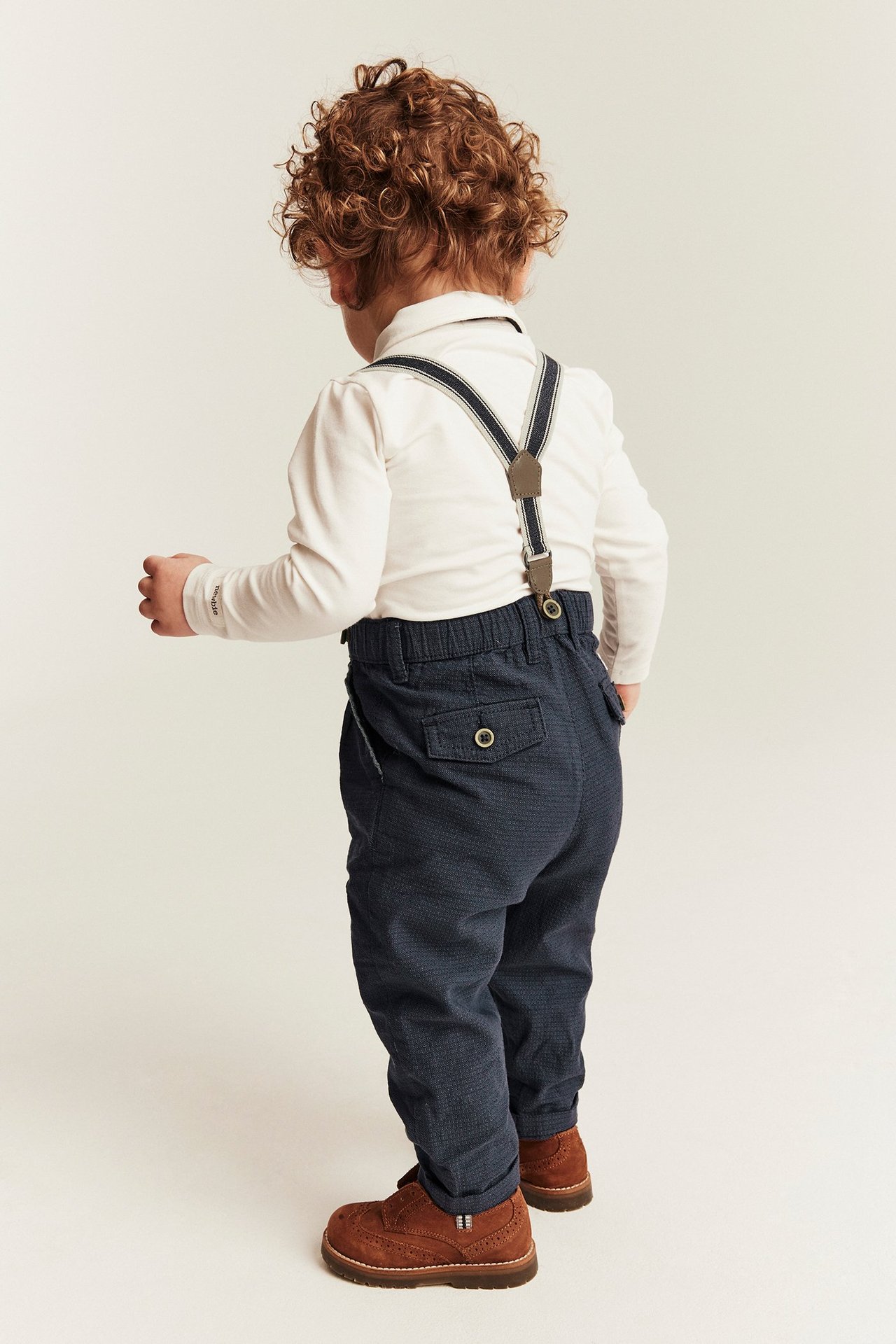 Back view: Kid in dark blue textured trousers with striped suspenders, white shirt, and brown boots.