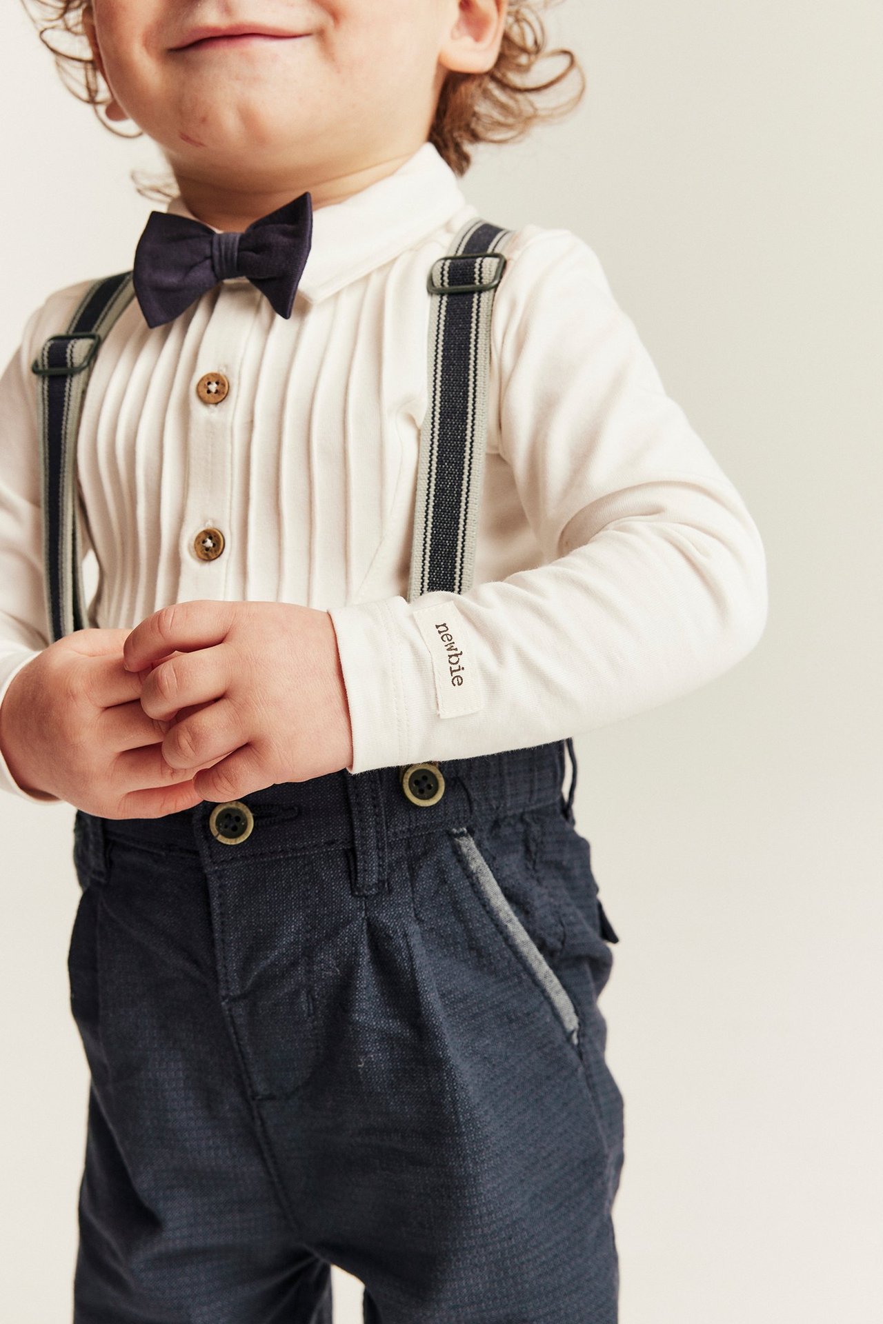 Kid wears dark blue woven dungaree trousers with striped suspenders, a white pleated shirt, and dark blue bow tie.