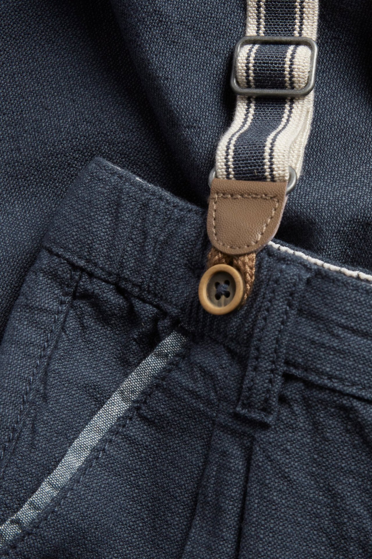 Nahaufnahme dunkelblauer Kinder-Latzhose mit braunem Knopf und gestreiftem Hosenträger.