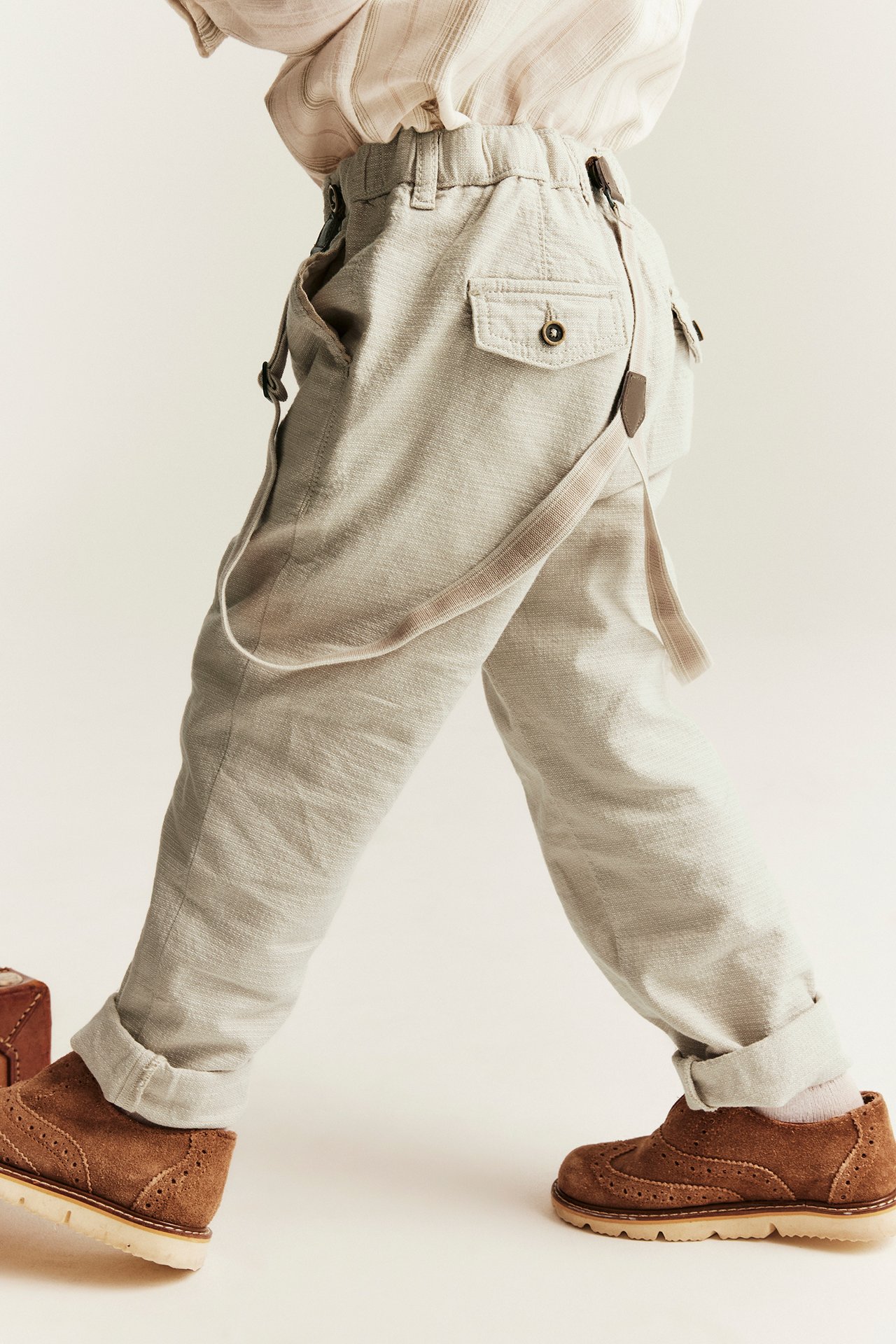 Side view of a kid in beige woven trousers with suspenders, a light striped shirt, and brown suede shoes.