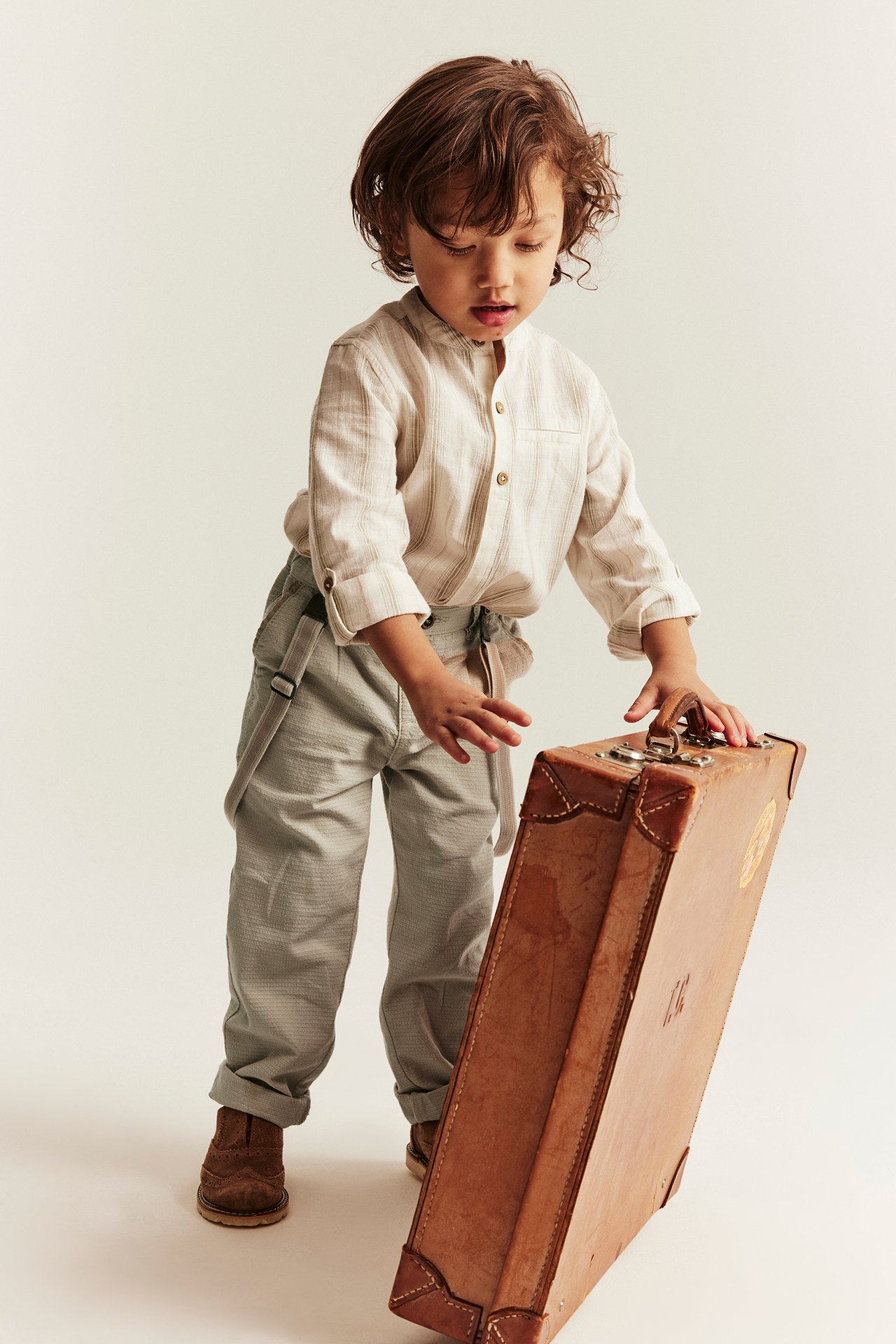 Front view: Kid wears light beige woven trousers with suspenders, a striped shirt, and brown boots, with hands on a suitcase.
