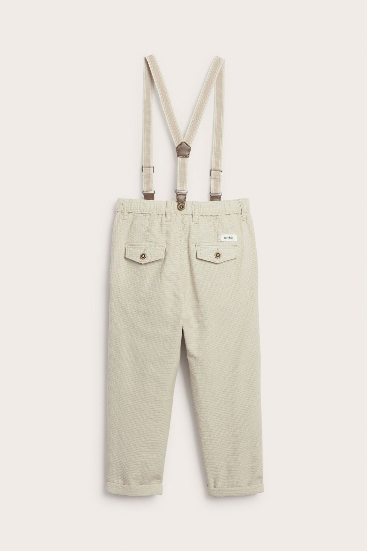 Rückenansicht: beige gewebte Babyhose mit verstellbaren Hosenträgern und Gesäßtaschen, mit aufgerollten Bündchen.