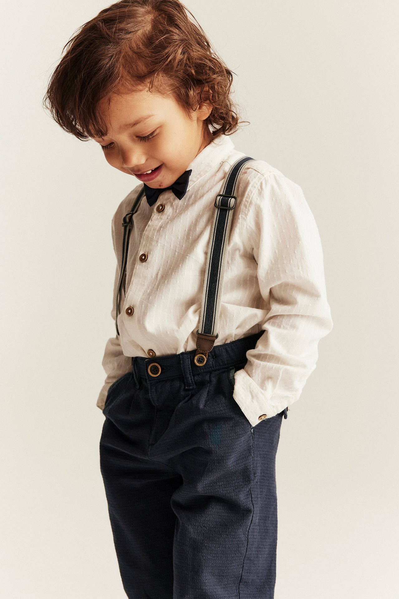 Front view: kid in dark blue woven suspender trousers, off-white shirt, and a dark blue bow tie.