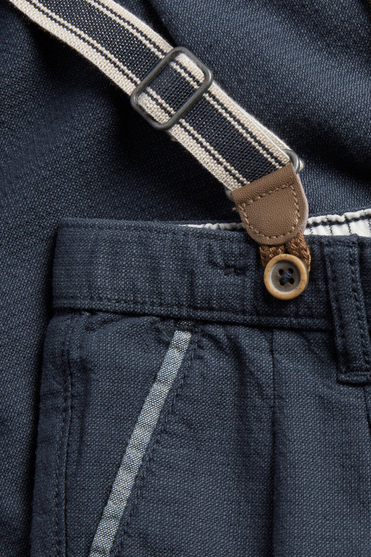 Close-up of dark blue woven cotton suspender trousers with a striped suspender, faux leather detail, and button.