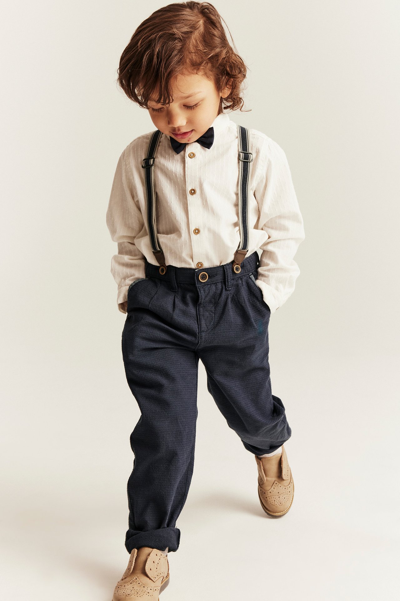 Kid in dark blue woven suspender trousers, light striped shirt, dark bow tie, and brown shoes. Front view.