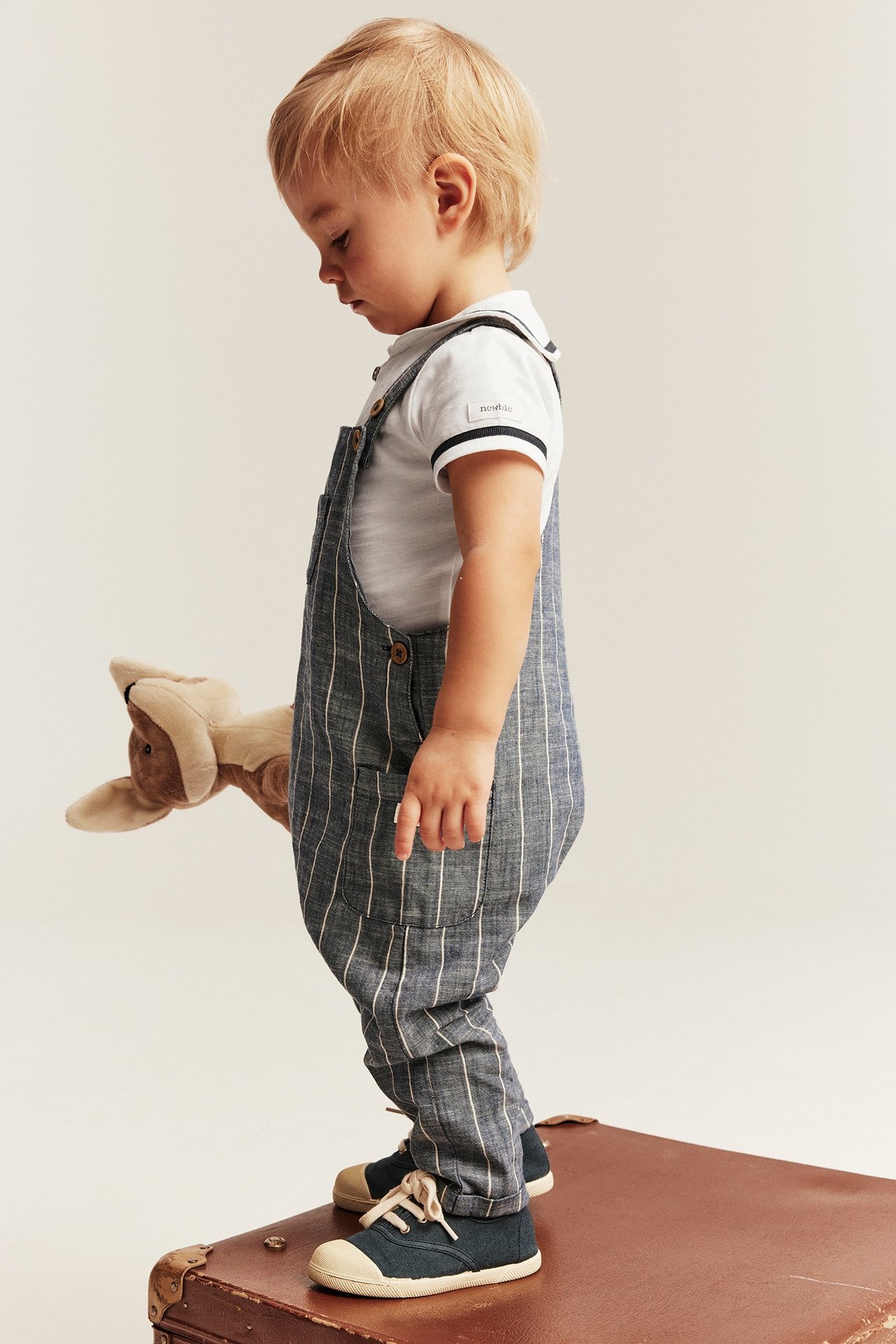 Seitenansicht eines Babys in offwhite Matrosenbody, blau gestreiften Overalls, dunkelblauen Stoffschuhen, hält Känguru-Spielzeug.