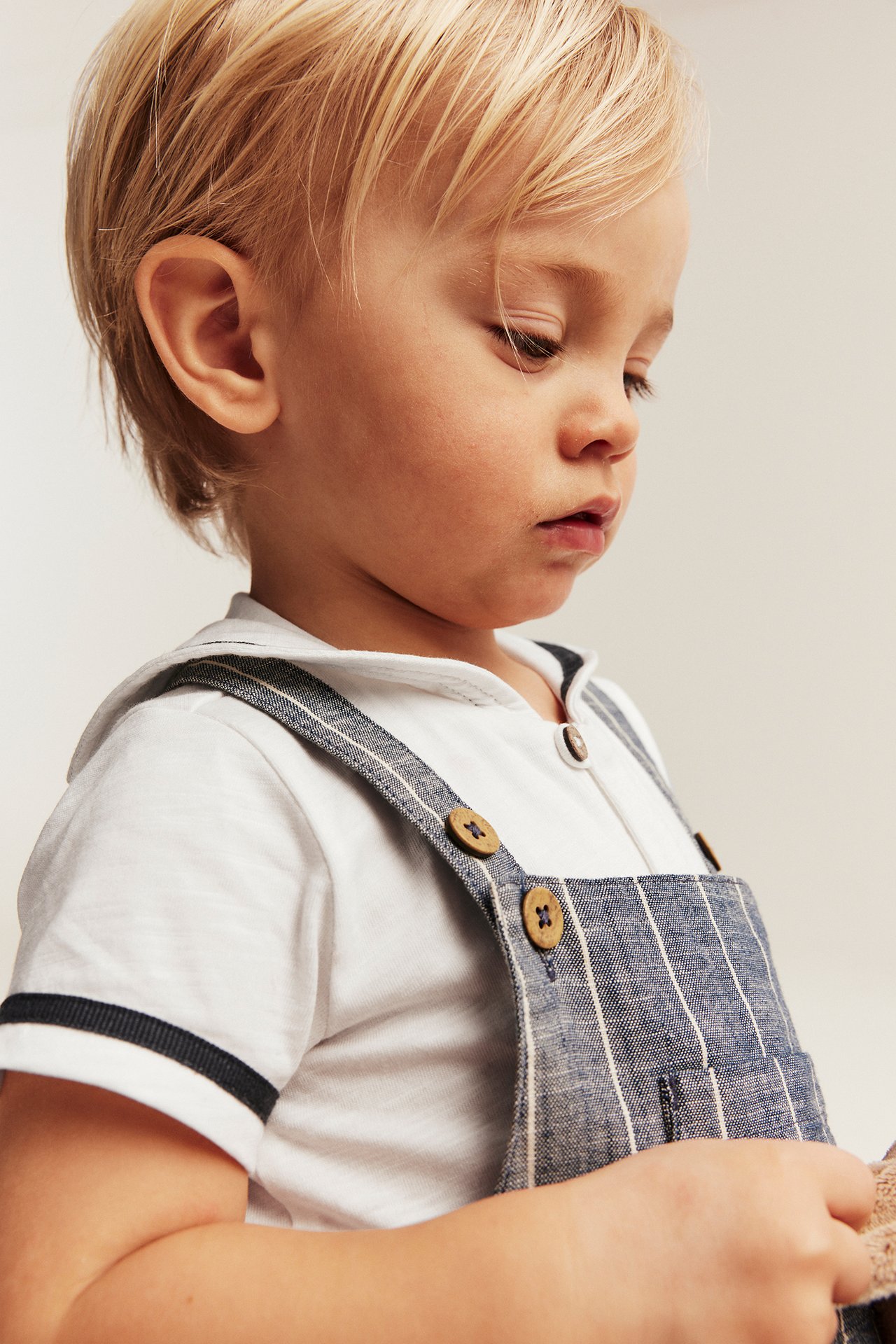 Close-up side view of a baby wearing an off-white short-sleeved sailor body layered under blue striped dungarees with wooden buttons.