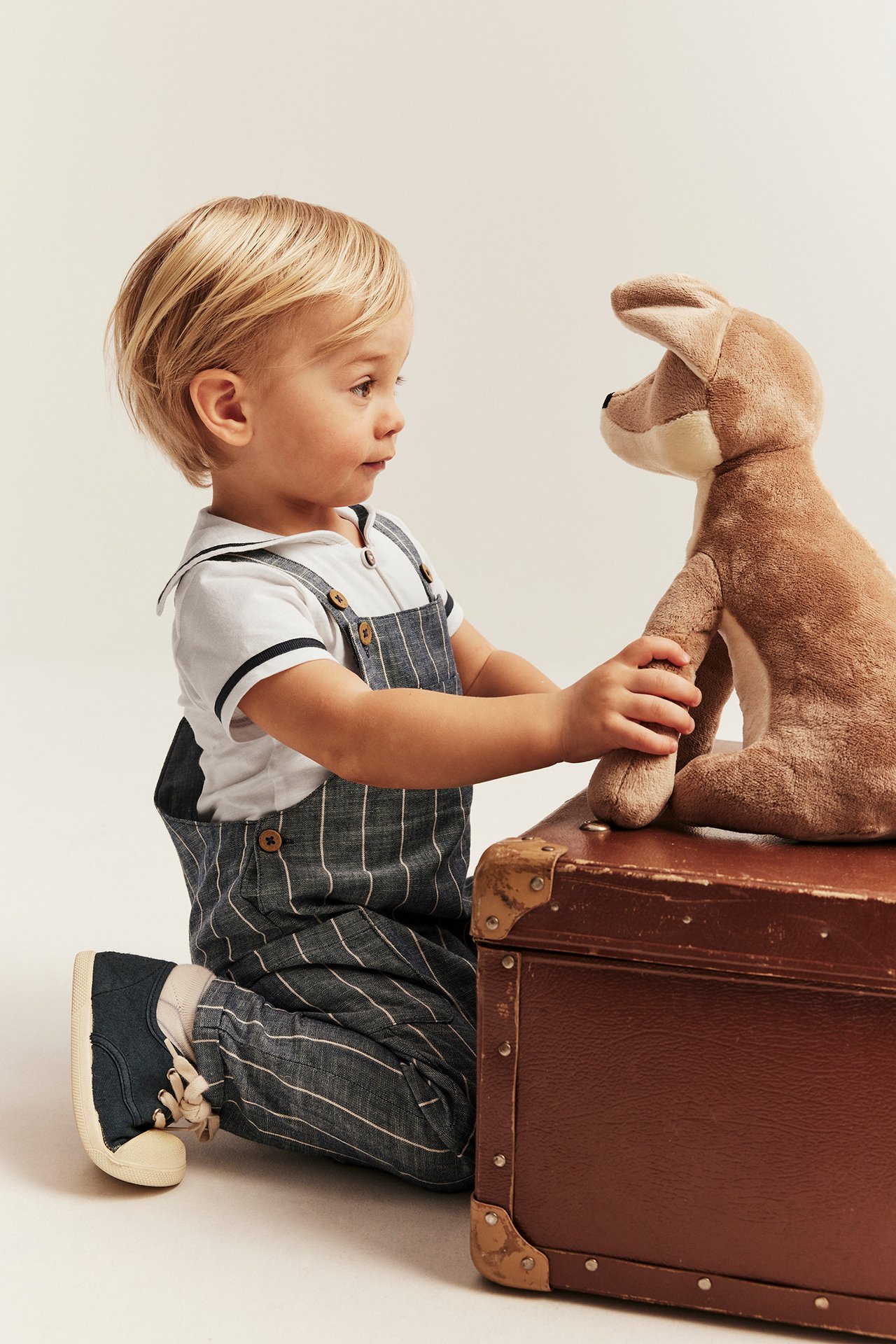 Seitenansicht: Baby in cremeweißem Matrosenbody, gestreiften Overalls, blauen Schuhen, hält ein braunes Plüschtier.