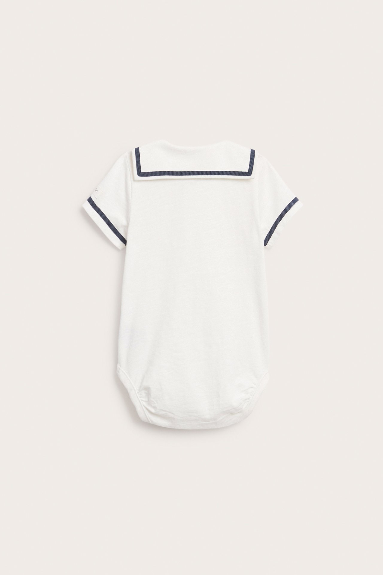 Back view of an off-white baby short-sleeved cotton bodysuit with a navy-trimmed sailor collar and sleeves.
