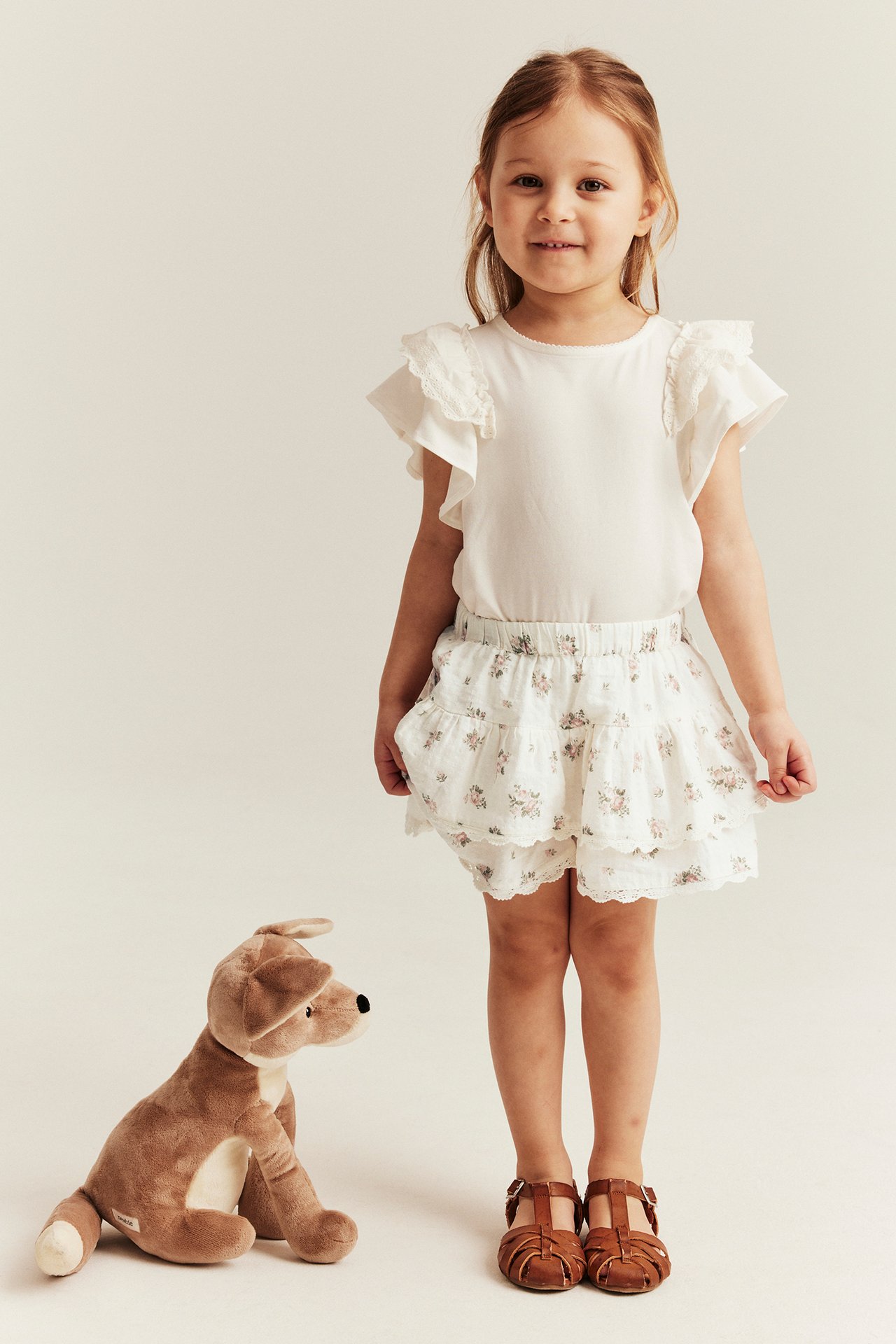 Vorderansicht: Kind in cremefarbenen geblümten Rock-Shorts, cremefarbenem Oberteil und braunen Sandalen.