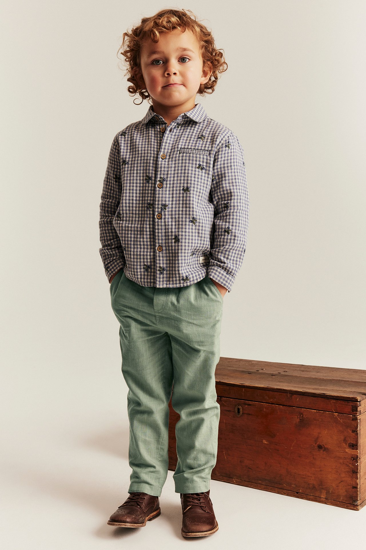 Front view of a kid wearing light green textured cotton trousers, a checked shirt, and brown lace-up boots.