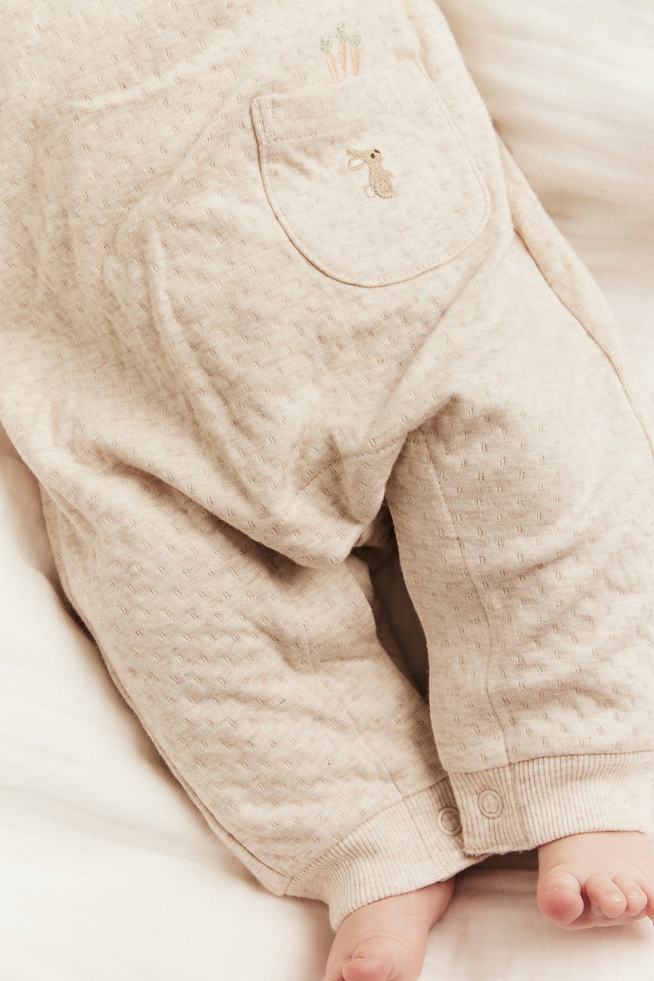 Nahaufnahme von Baby in beiger Melange Jersey-Latzhose mit gestickter Kaninchen-Tasche und Rippbündchen.