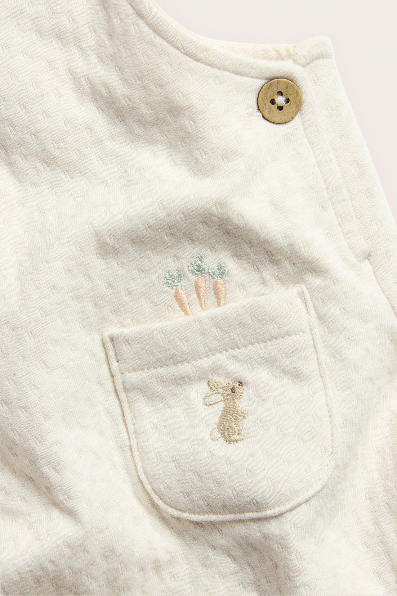 Close-up of a beige melange baby jersey overall with a front pocket featuring bunny and carrot embroidery.
