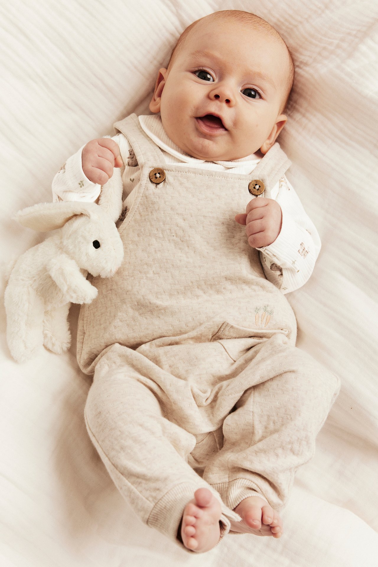 Front view: Baby in beige jersey dungarees with carrot embroidery over a white top, holding a white bunny.