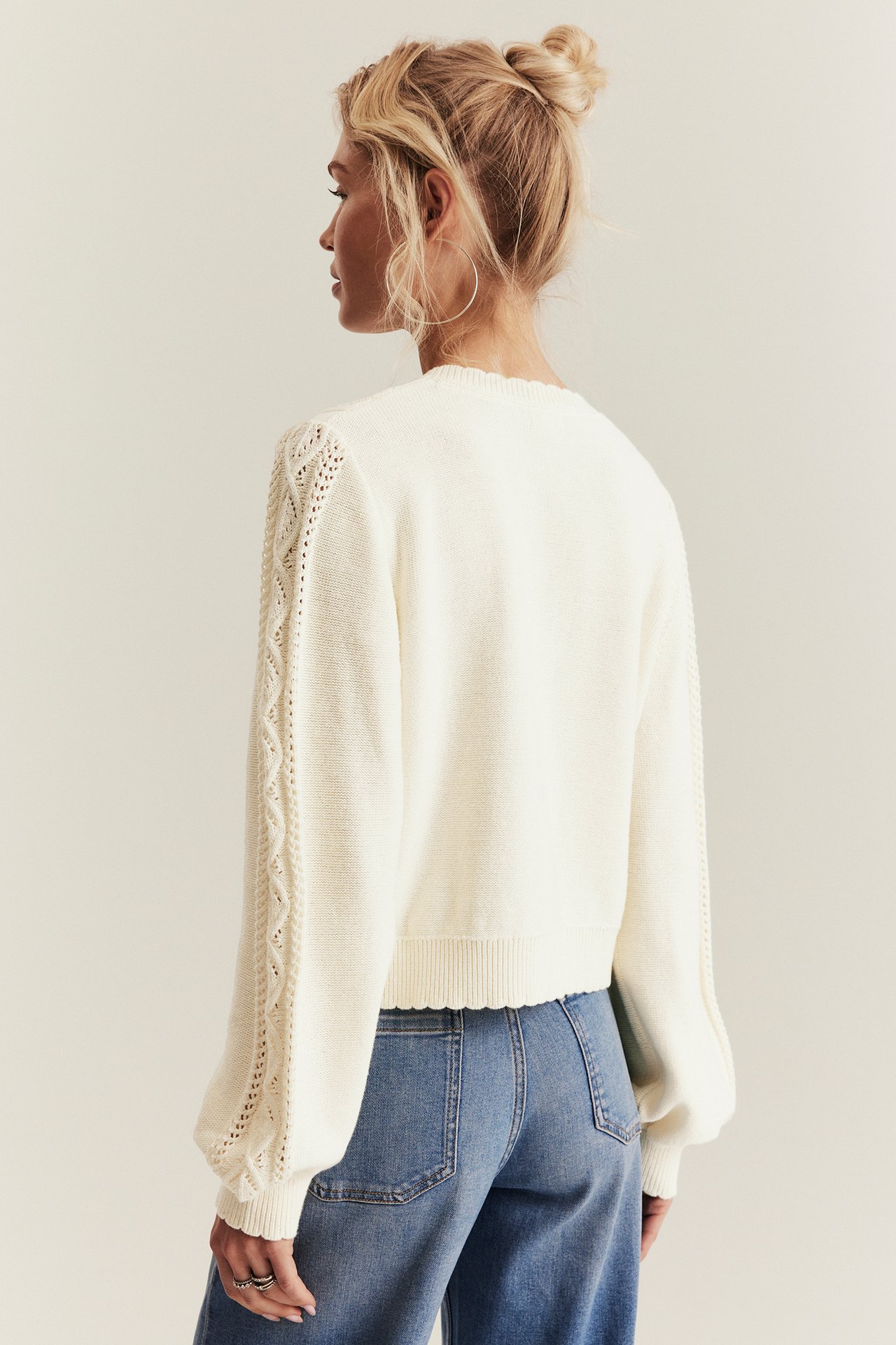 Woman's back view in an off-white knitted top with openwork patterned sleeves and blue jeans.