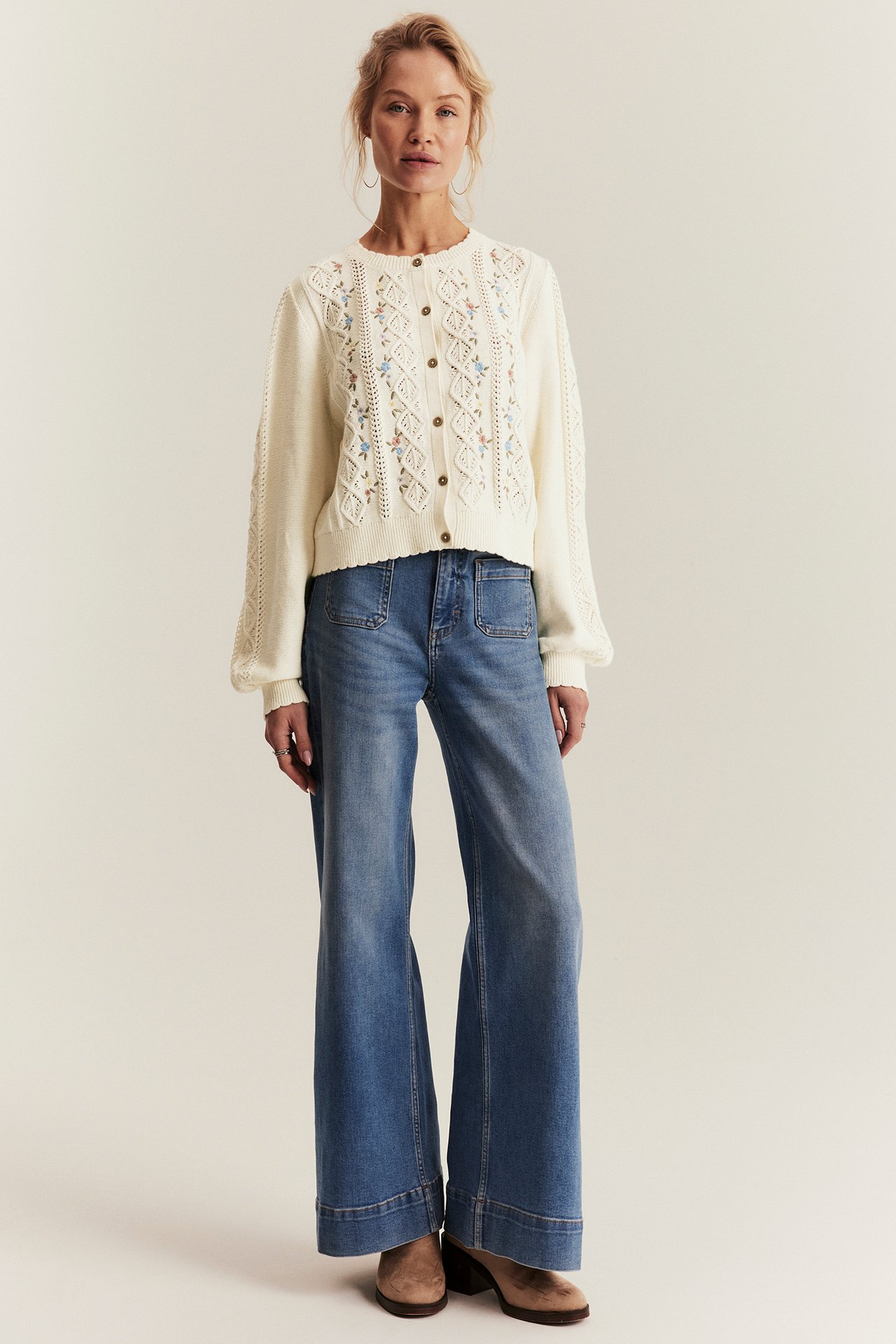 Front view of a woman wearing an offwhite cotton embroidered cardigan, blue wide-leg jeans, and brown boots.