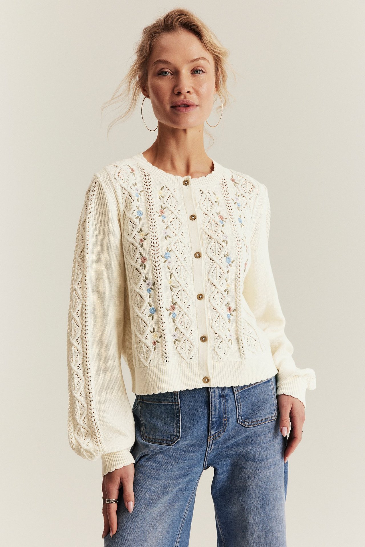 Front view of a woman wearing an off-white cotton knitted cardigan with floral embroidery, buttons, and blue jeans.